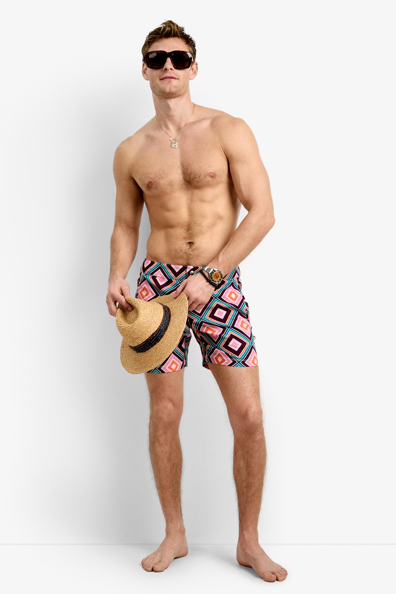 A shirtless man wearing Honolulu Board Short swim trunks with colorful geometric patterns, dark sunglasses, a wristwatch, and a necklace stands barefoot against a white background, holding a straw hat by his side.