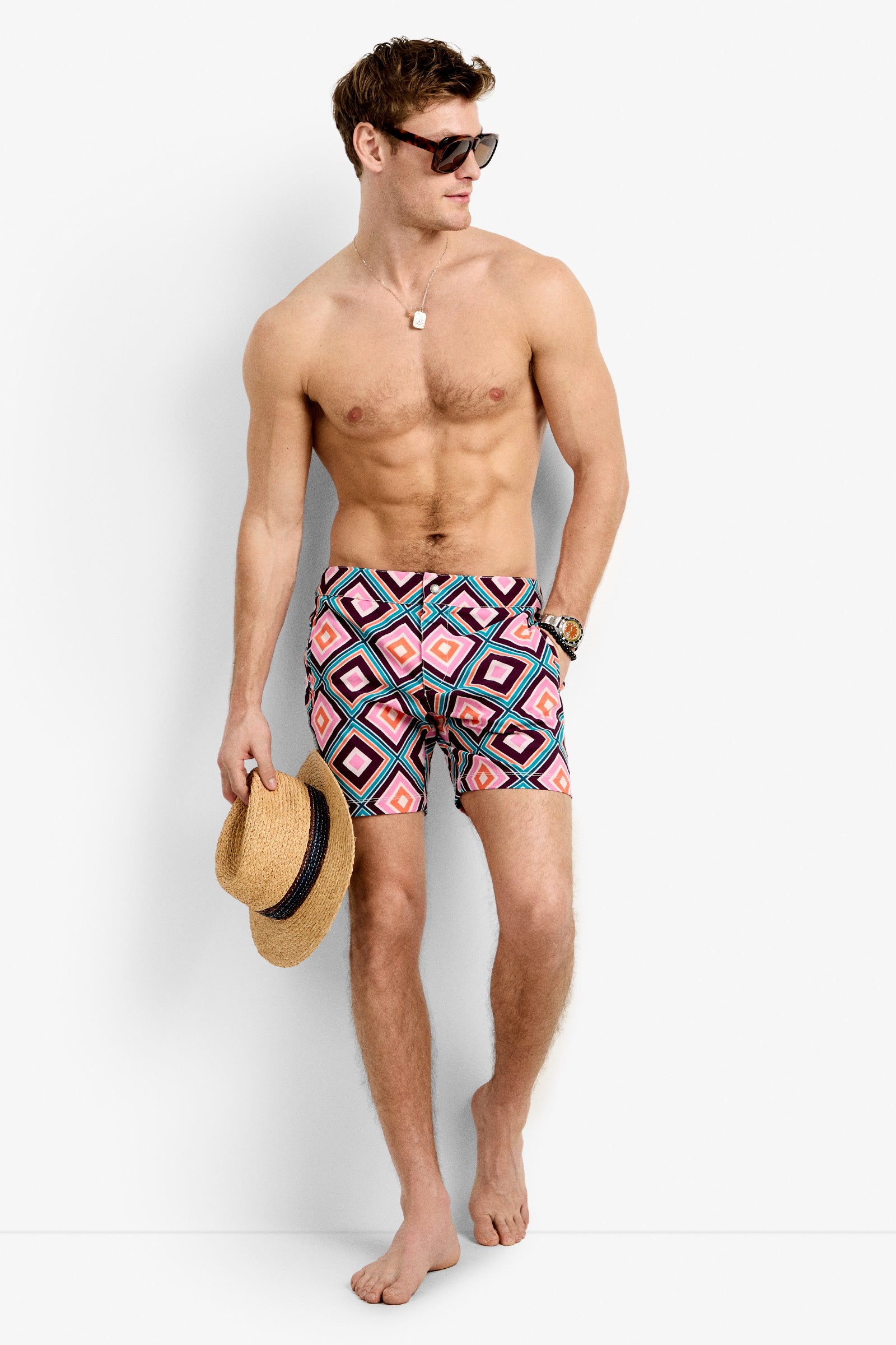 A shirtless man stands barefoot against a white background, wearing Honolulu Board Shorts with colorful geometric patterns and sunglasses, holding a straw hat in his left hand while looking to the side.