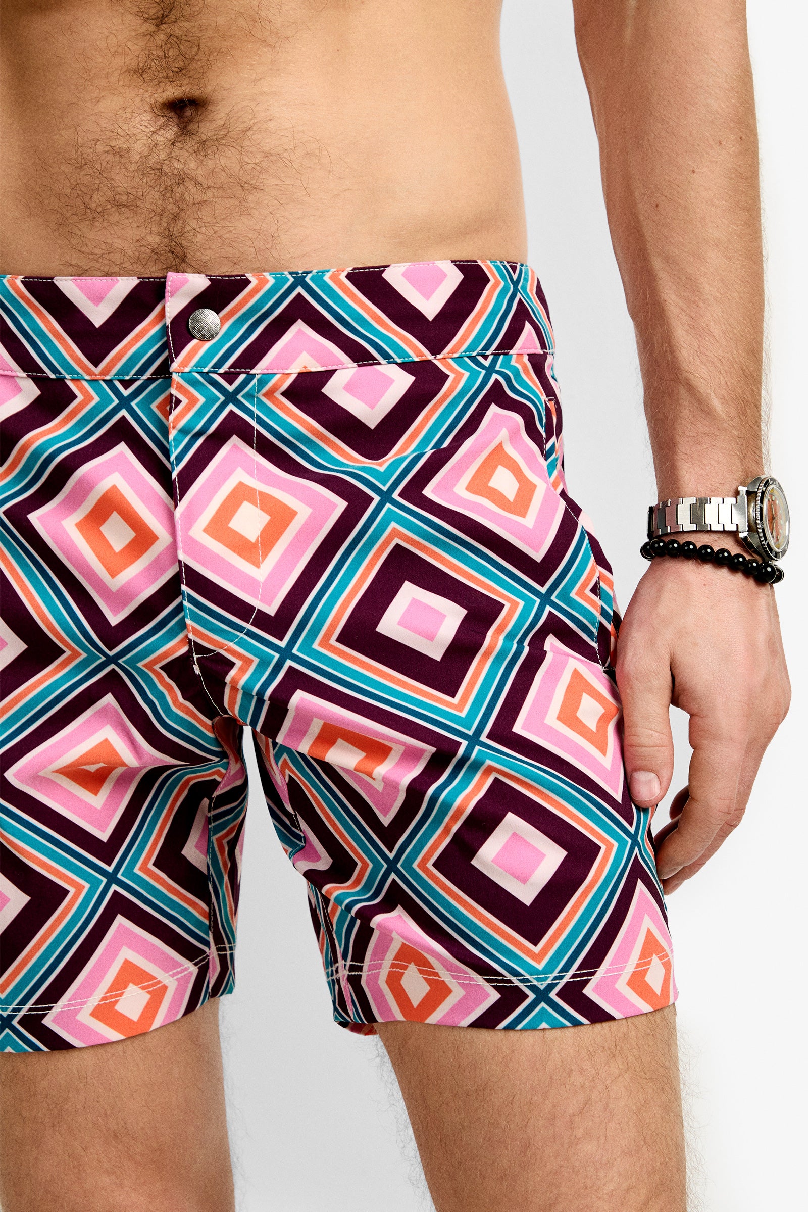 A person wears the Honolulu Board Short featuring colorful geometric diamond patterns in pink, orange, purple, and blue. Their torso is bare, and they have a wristwatch and bracelet on their left wrist.