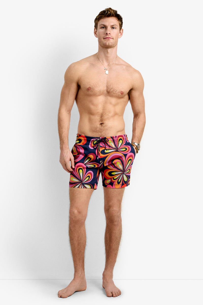 A barefoot man with short light brown hair stands facing forward, shirtless, wearing the Imperial Beach Swim Trunk - Boca Blooms featuring a bold psychedelic pattern in pink, orange, yellow, and black.