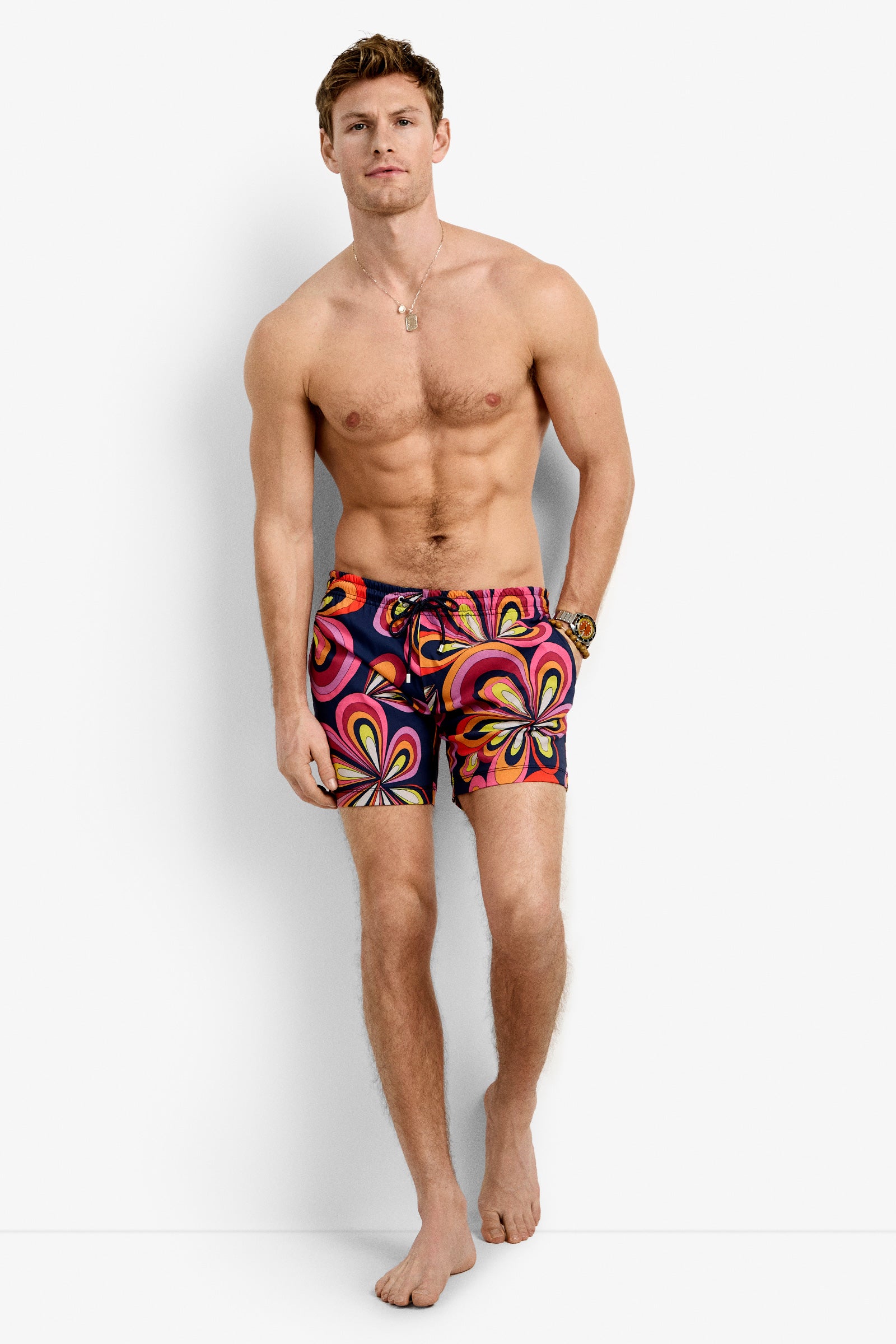 A shirtless, barefoot man with short brown hair stands against a white background, wearing the Imperial Beach Swim Trunk - Boca Blooms featuring bold, bright swirling floral patterns in red, pink, yellow, and orange.