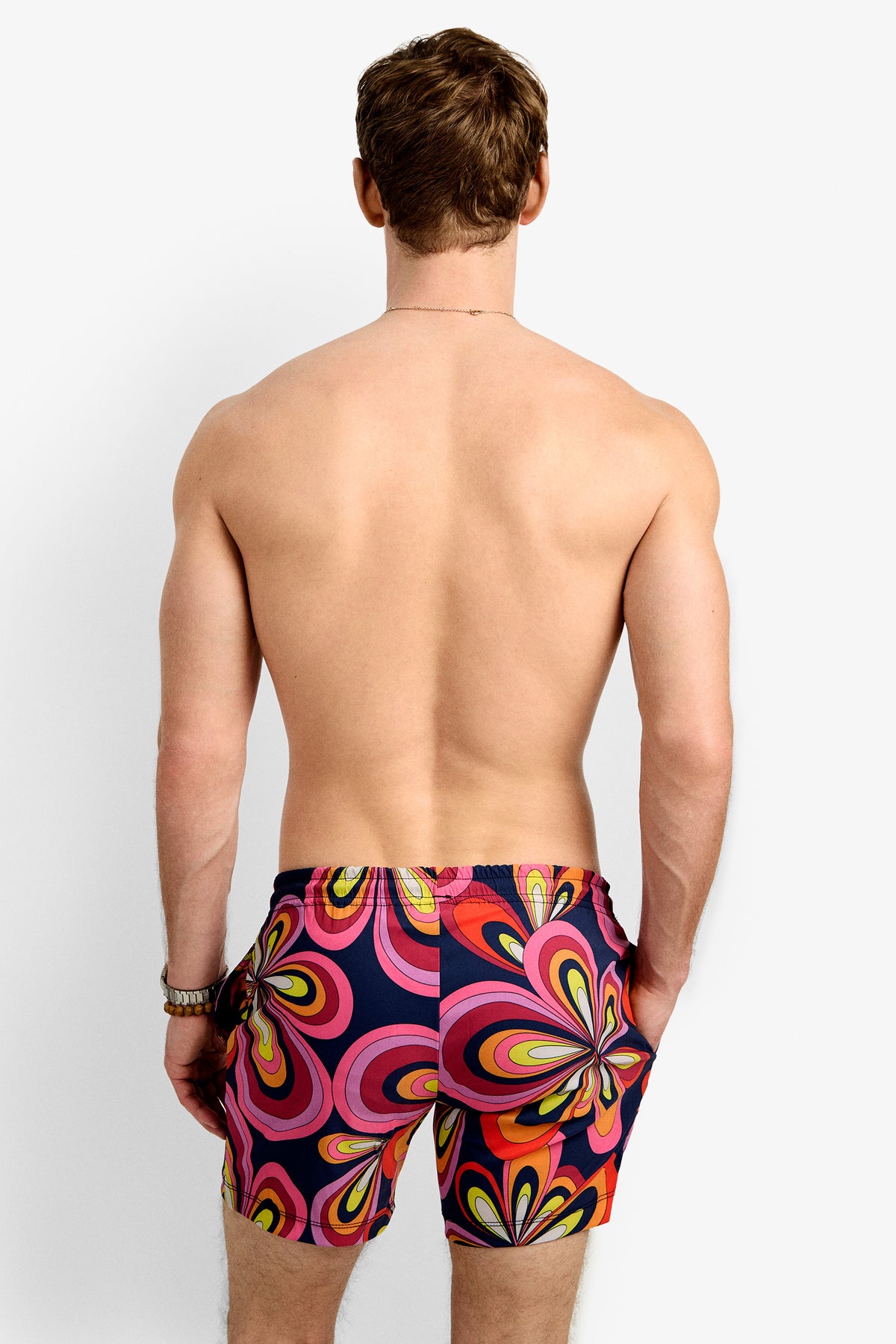 A man with short brown hair, shirtless and facing away, wears the Imperial Beach Swim Trunk - Boca Blooms featuring a bold, retro floral pattern in pink, yellow, orange, and navy blue.