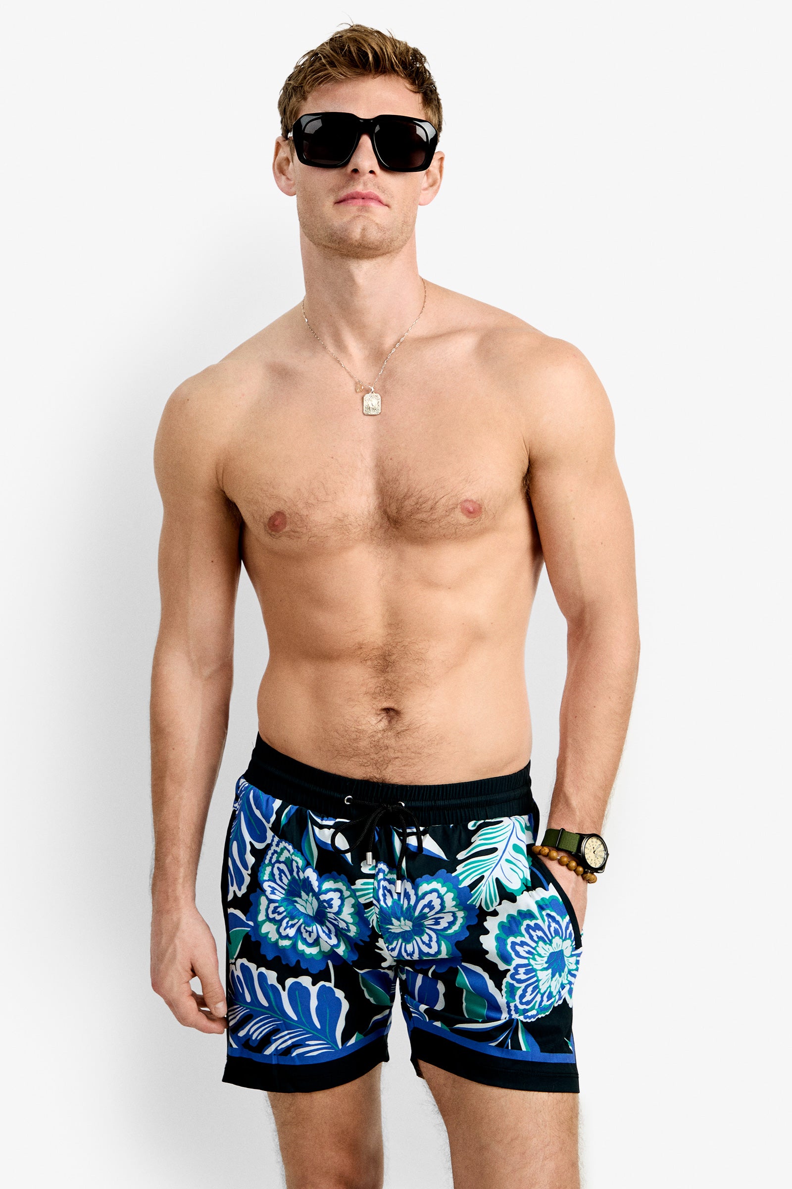 A shirtless man with short light brown hair, dark sunglasses, a gold necklace and watch models Bruce's Beach Swim Trunk—floral blue swim trunks—against a plain white background.