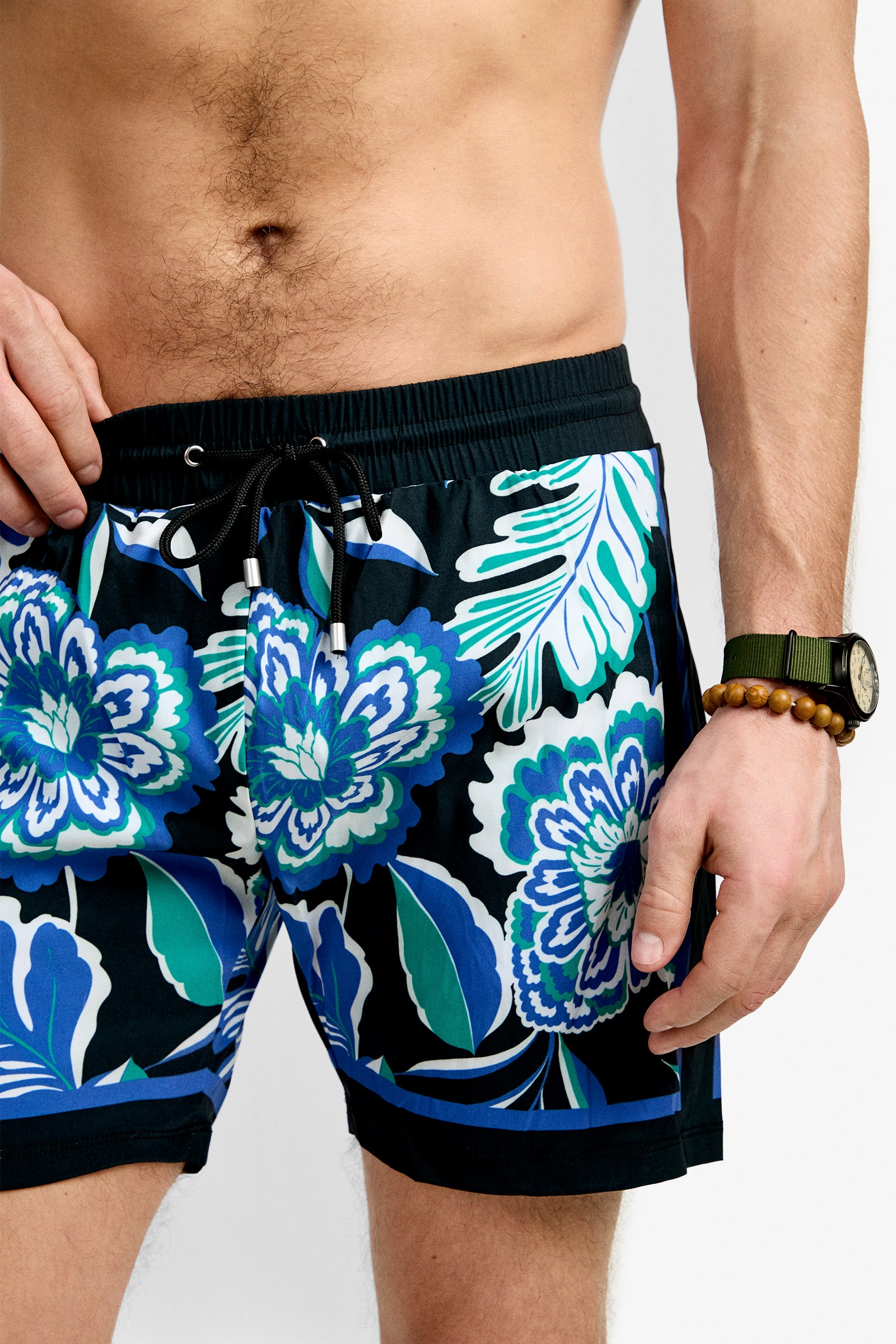 A man wears Bruce's Beach Swim Trunk, featuring black fabric with blue, green, and white floral patterns. A beaded bracelet and a wristwatch adorn his left wrist. His torso and mid-thighs are visible.