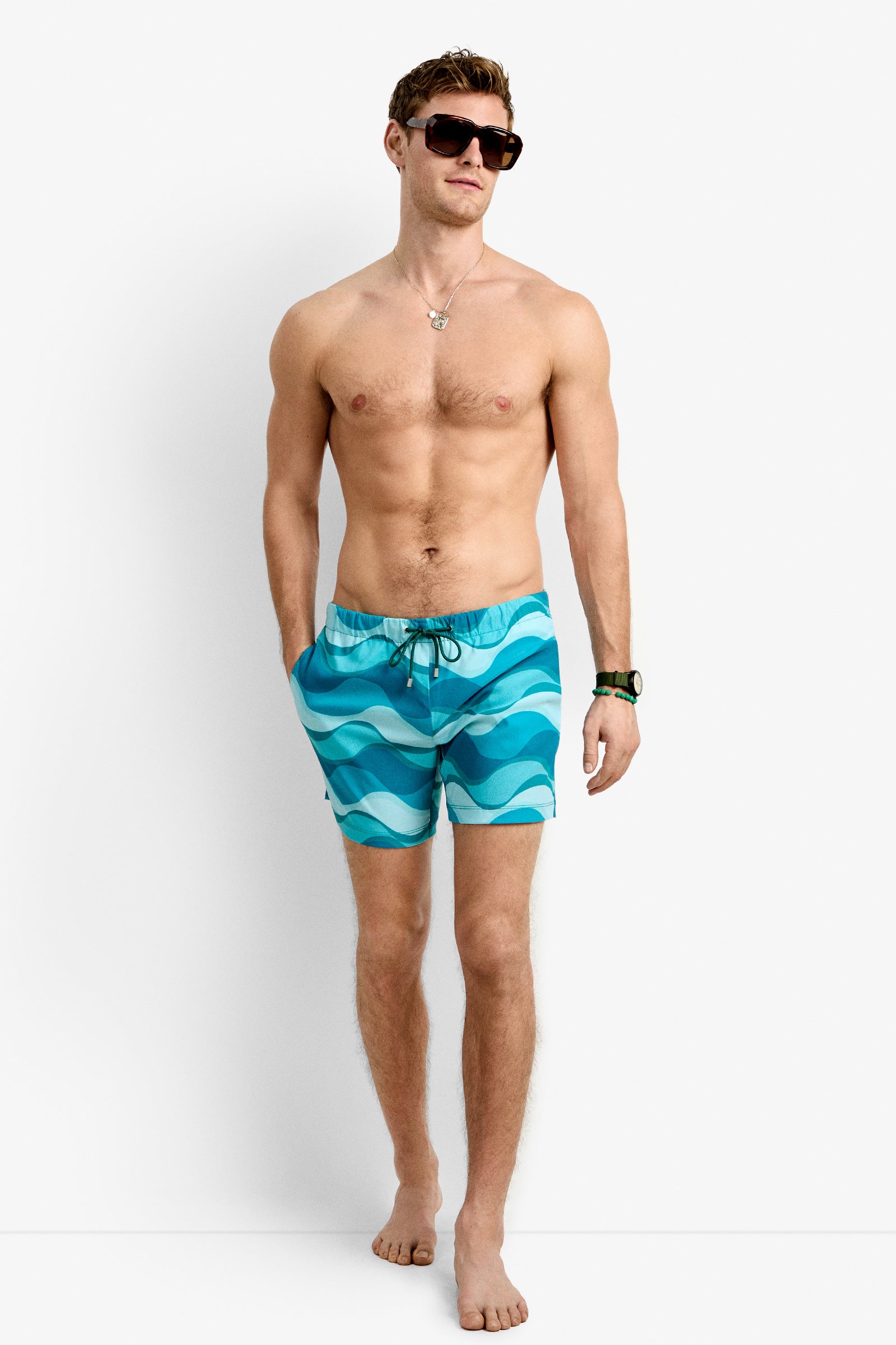 A shirtless man wearing sunglasses, a necklace, a watch, and Imperial Beach Swim Trunk in blue stands barefoot with his hands in his pockets against a plain white background.