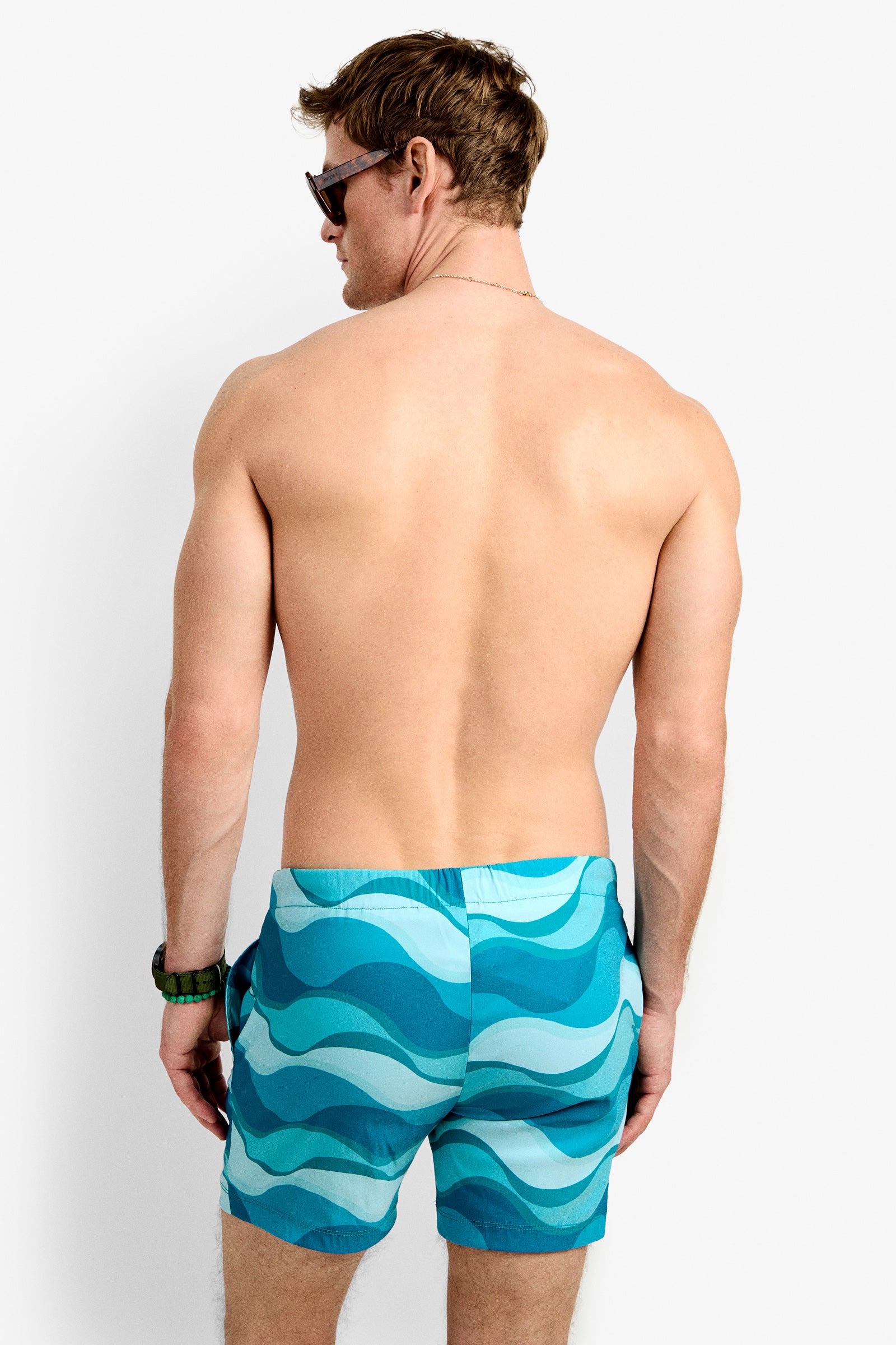 A shirtless man with short brown hair, sunglasses, and a bracelet stands with his back to the camera, wearing Imperial Beach Swim Trunks featuring a blue and teal wavy pattern.