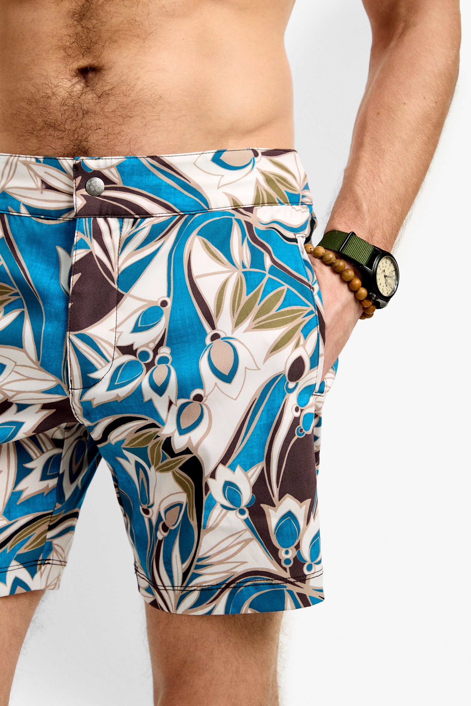 A man wears the Honolulu Board Short featuring blue, brown, and white tropical patterns. He accessorizes with a green wristwatch and wooden beaded bracelet on his left wrist, his hand tucked into the shorts pocket.