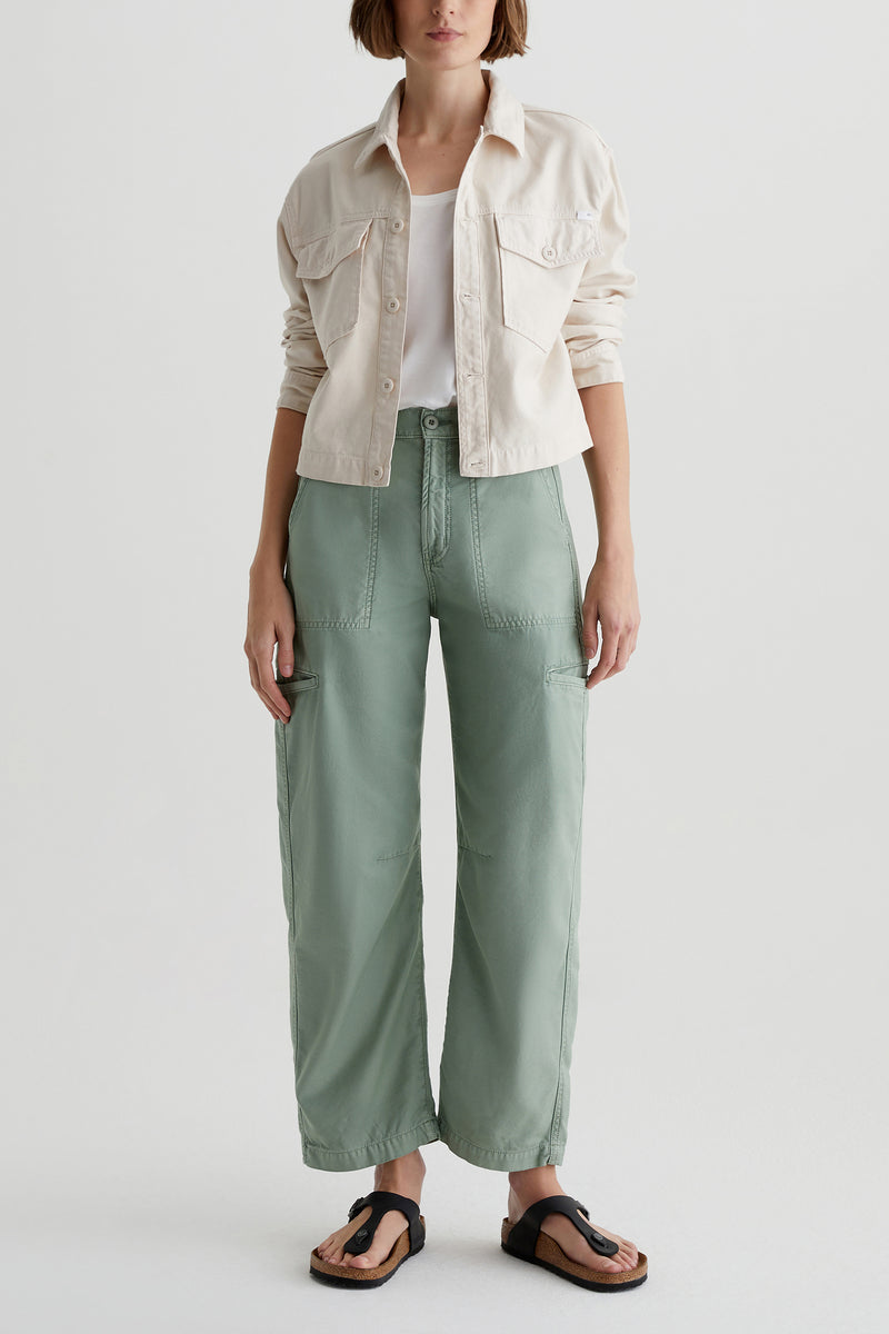 A person stands against a plain background wearing a light beige jacket, white top, and sage green AG Hazal High-Rise Utility Barrel pants with large pockets. Black sandals complete the look; their face is partially out of frame.