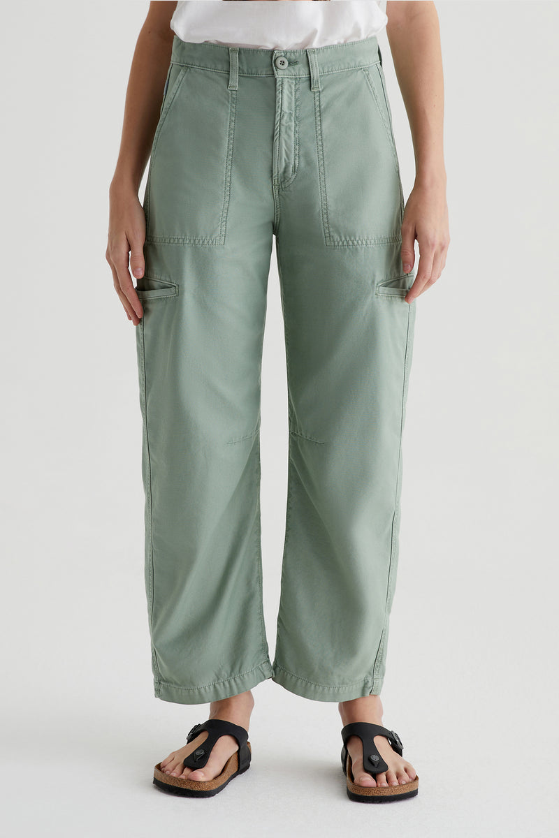 A person wearing AG Hazal High-Rise Utility Barrel pants in Matcha with front patch pockets and a white shirt, standing in black sandals against a plain light background.