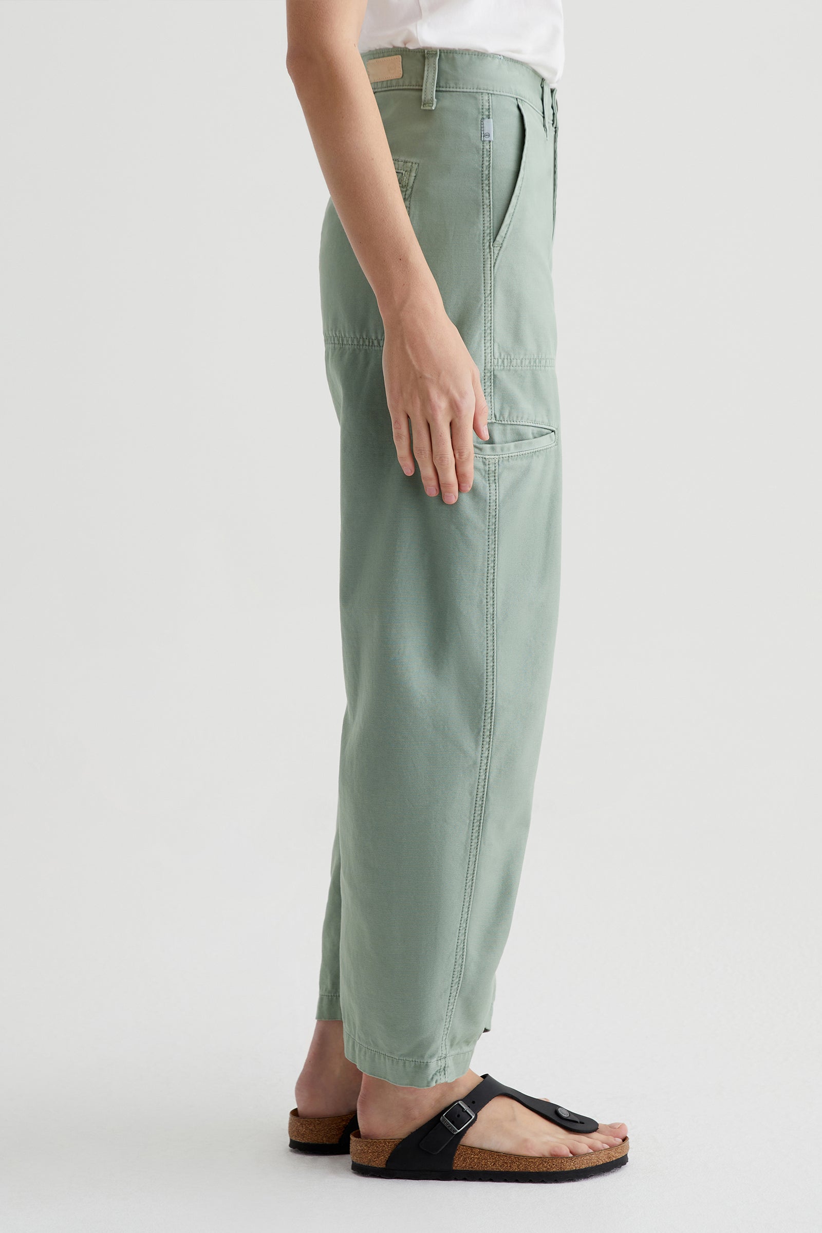 A person stands sideways against a plain light background, wearing the AG Hazal High-Rise Utility Barrel in sage green, paired with a white shirt and black sandals with cork soles.