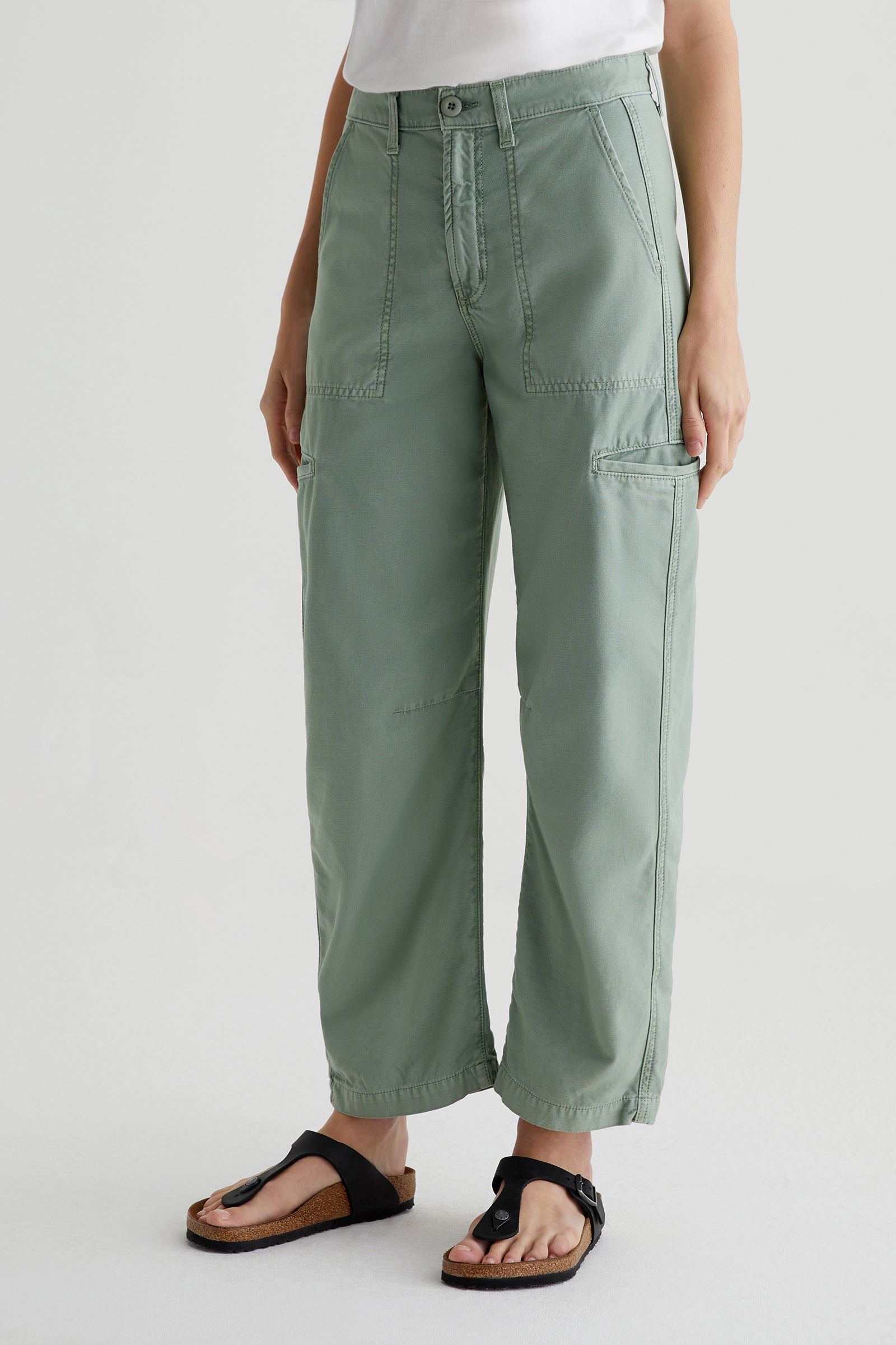 A person in AG Hazal High-Rise Utility Barrel sage green cargo pants with front and side pockets, black sandals, and a white shirt stands against a plain light background.