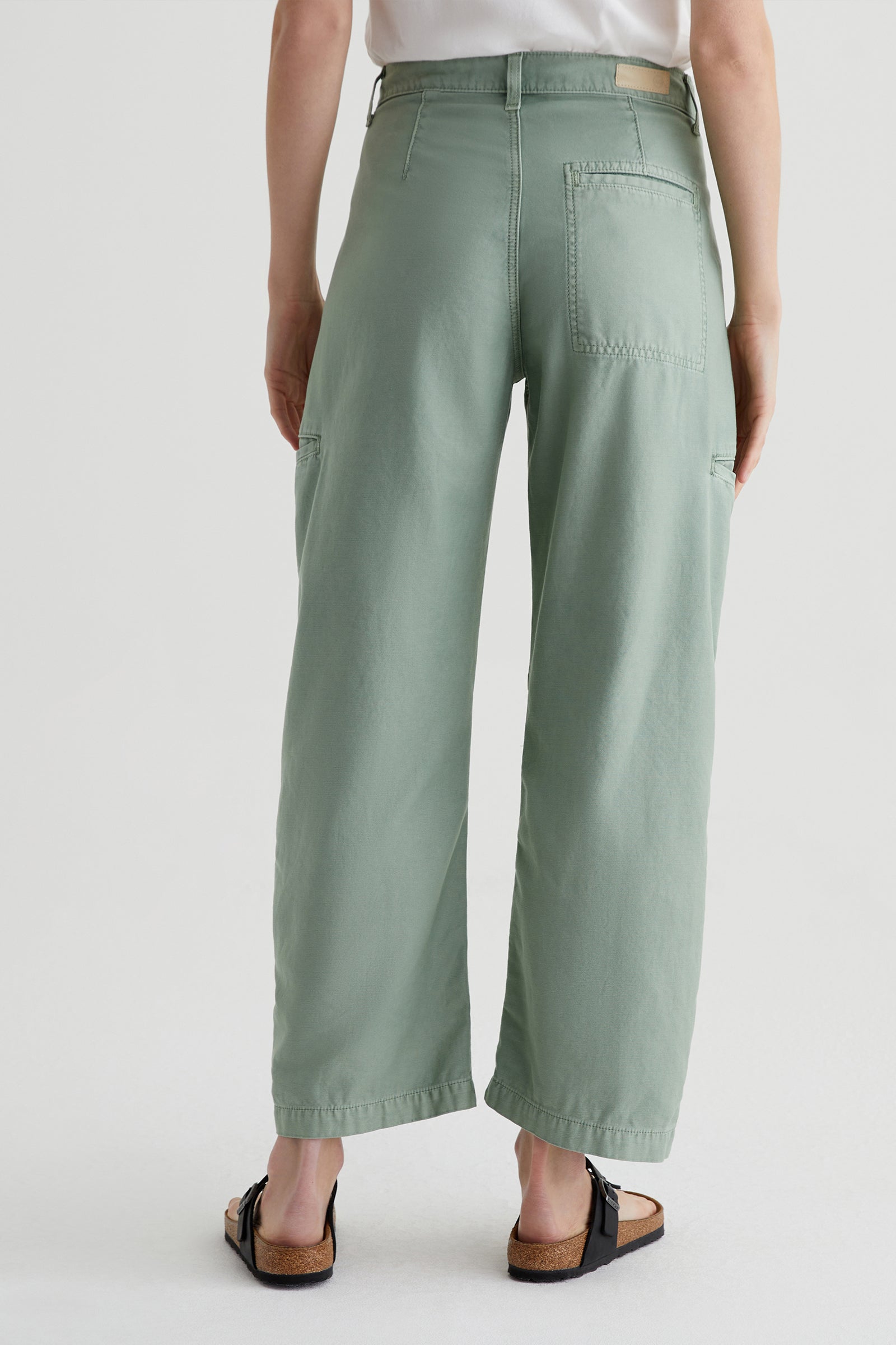 Someone wearing AG Hazal High-Rise Utility Barrel pants in sage green and black sandals stands with their back to the camera against a plain light background.