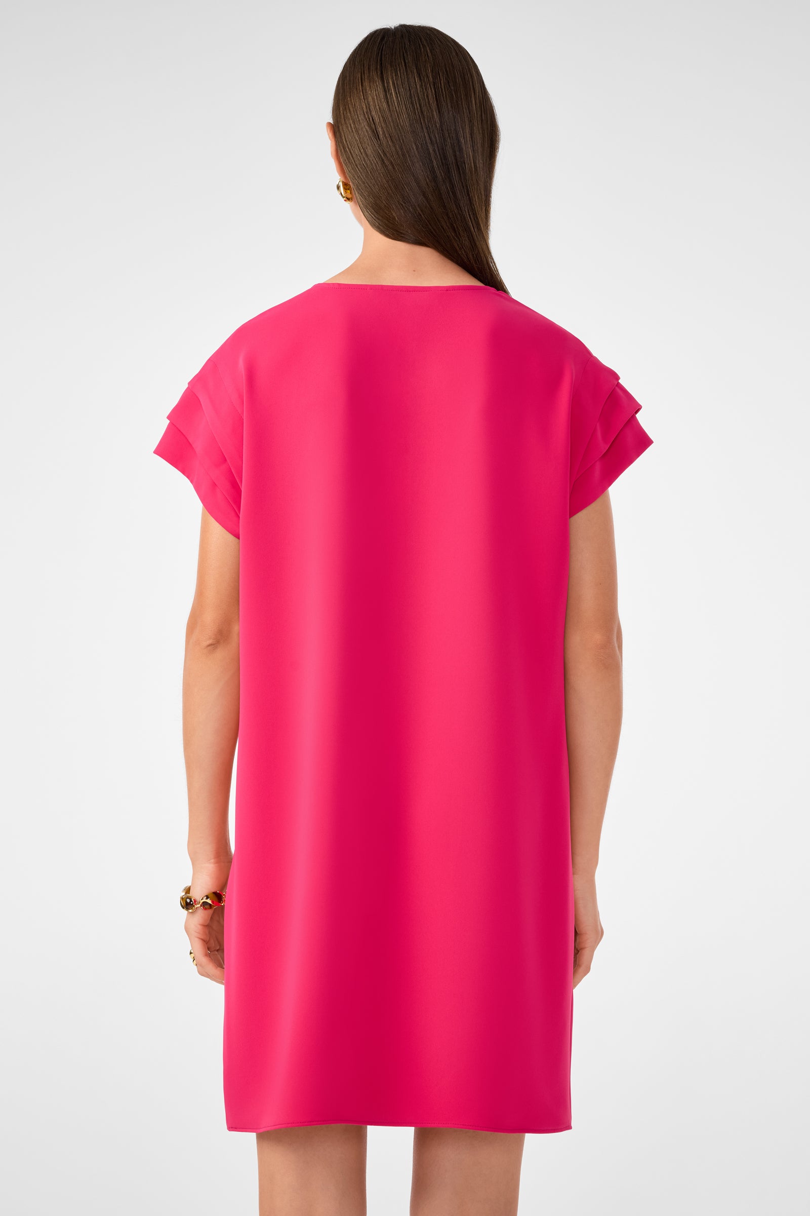A woman with straight brown hair, shown from the back against a light gray background, wears gold earrings, a bracelet, and the bright pink Sadora Short-Sleeve Dress.