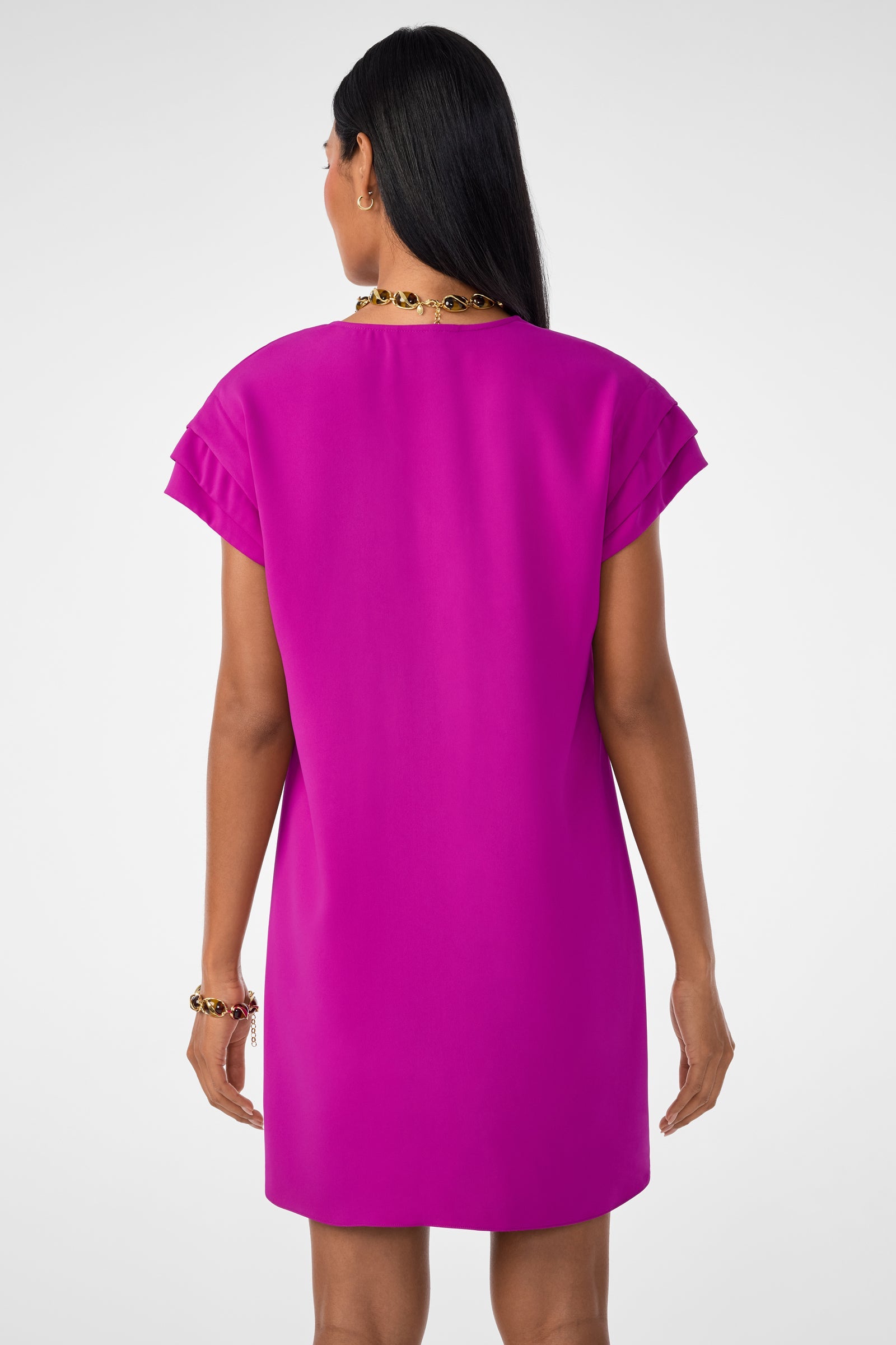 A woman with long dark hair is seen from the back wearing the Sadora Short-Sleeve Dress in geranium, accessorized with a gold necklace and bracelet, against a plain white background.