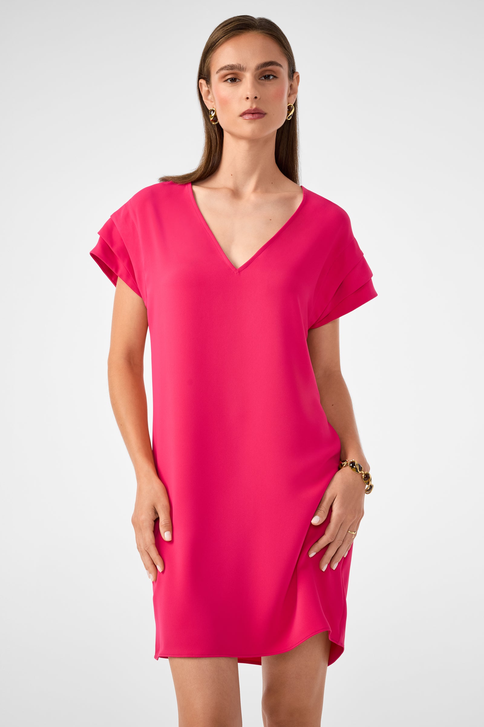 A woman with long brown hair wears the Sadora Short-Sleeve Dress in hot pink, featuring a V-neck and short sleeves. She accessorizes with gold earrings and a bracelet, standing against a plain light gray background.