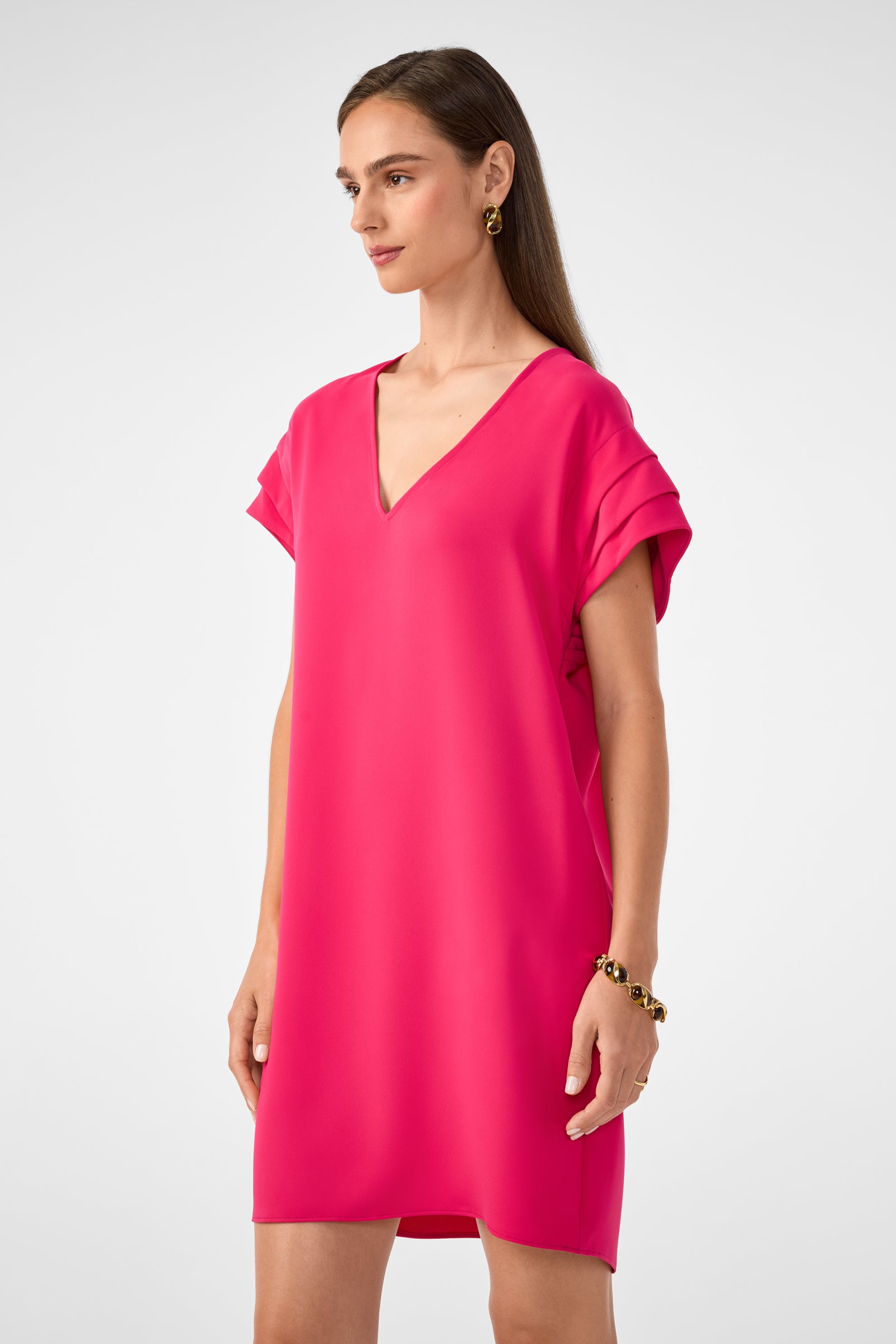 A woman with straight brown hair wears the Sadora Short-Sleeve Dress, a loose-fitting bright pink V-neck style, paired with gold earrings and a bracelet, standing against a plain light background.