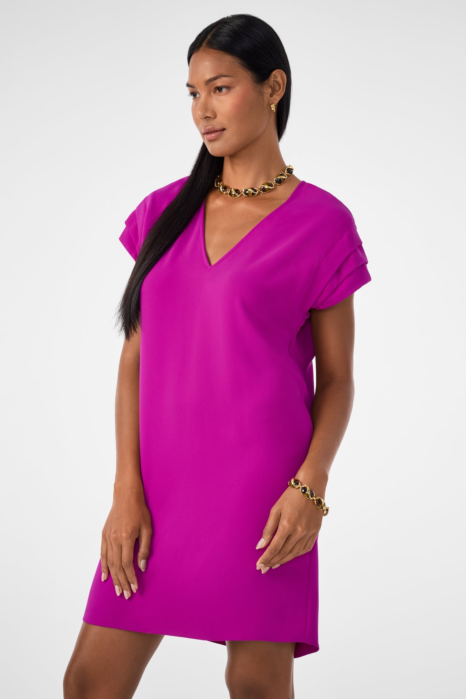 A woman with long black hair wears the Sadora Short-Sleeve Dress in Geranium, accessorized with a chunky gold necklace and bracelet, standing against a plain white background.