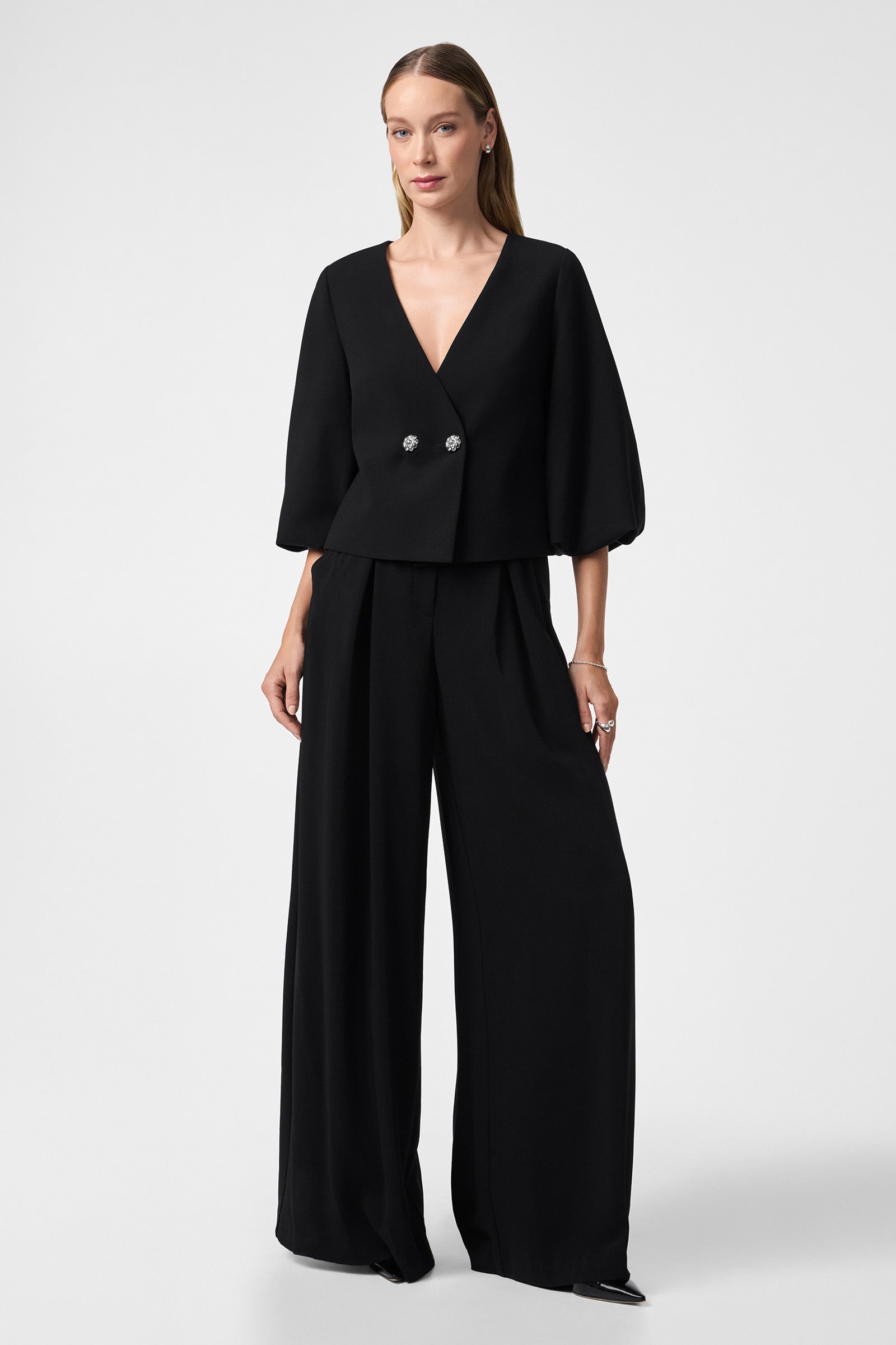 A woman stands against a plain background wearing a stylish black V-neck, double-breasted top with puffed three-quarter sleeves and Saige Wide-Leg Short Pants.