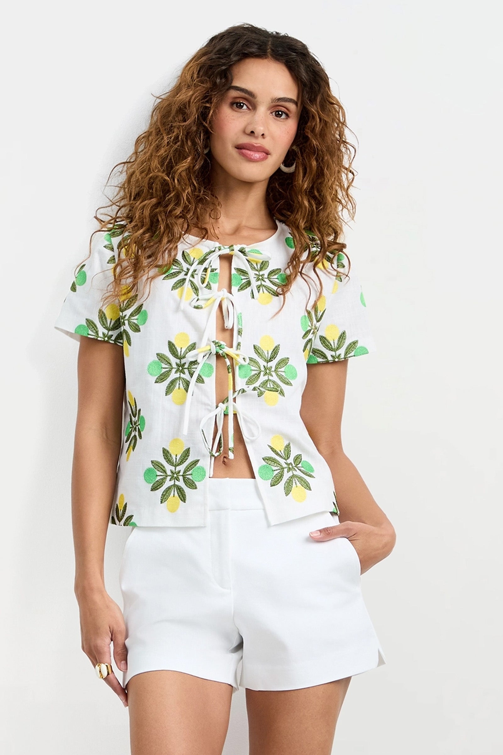 A woman with curly hair wears the Salina Tie-Front Top, a white short-sleeve top with green and yellow floral patterns and front ties, paired with white shorts. She stands against a plain white background with one hand in her pocket.
