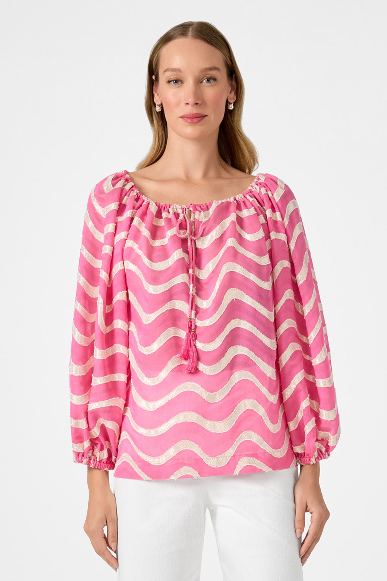 A woman wearing the Sandia 2 Blouson Top - Pinku, featuring white wavy stripes, long sleeves, and a gathered neckline with tassel ties, paired with white pants, stands against a plain light background.