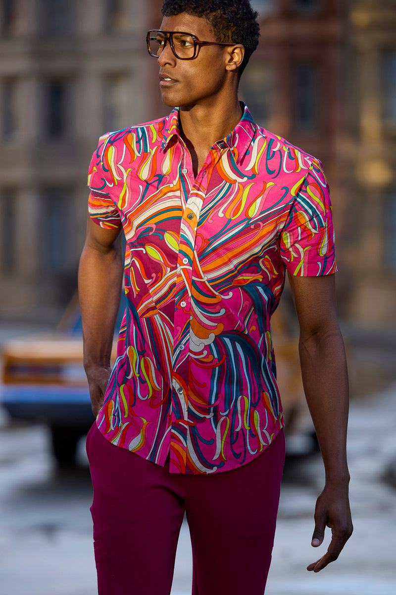 A man in glasses wears the Slim Jim Shirt, a slim-fit short-sleeve button-up with swirling pink, orange, yellow, and blue patterns, paired with magenta pants. He stands outdoors with blurred buildings and a car behind him.