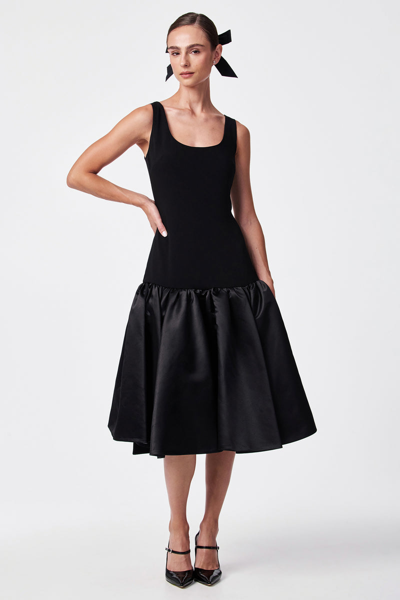 A woman models the SINA DRESS - BLACK, featuring a sleeveless fitted bodice and flared satin skirt. She accessorizes with a black ribbon in her hair and black heels against a plain white background.