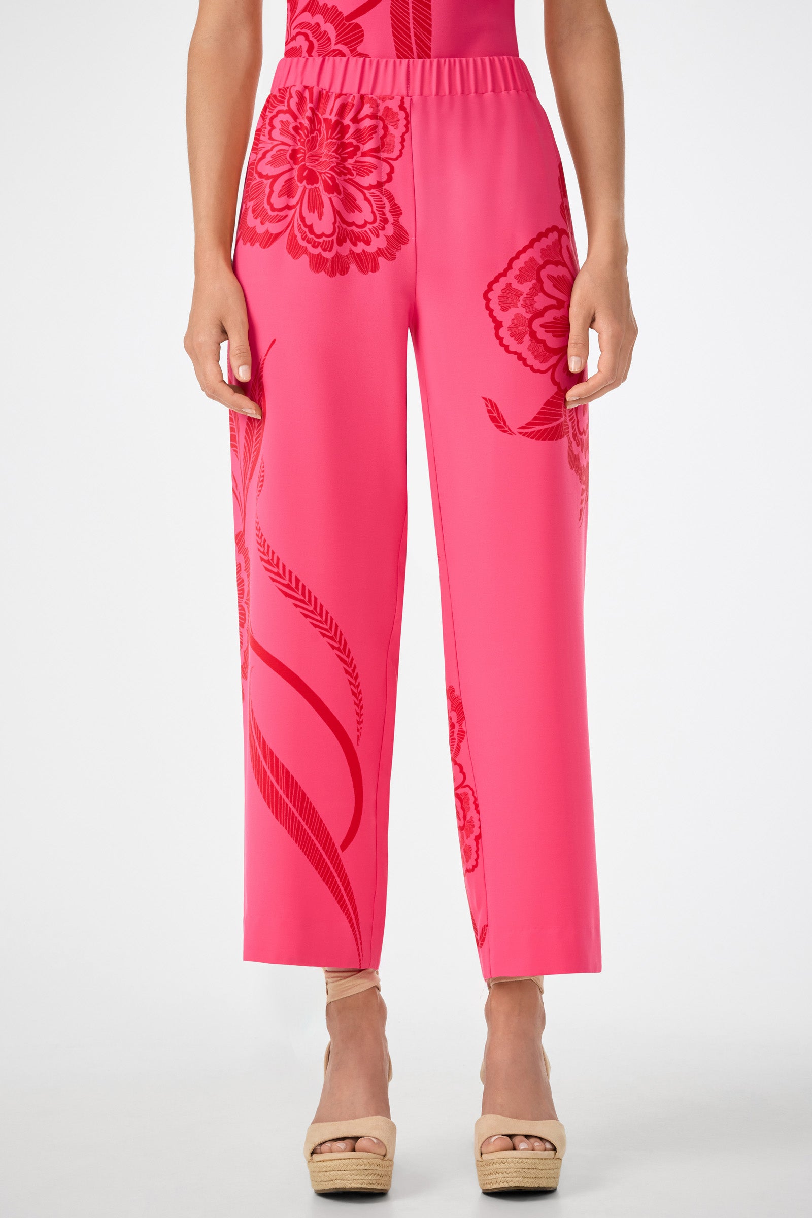 Model wears the Statue Pant—bright pink wide-leg trousers with bold red floral patterns—paired with beige wedge sandals. Cropped photo shows only the lower half of the body against a plain background.