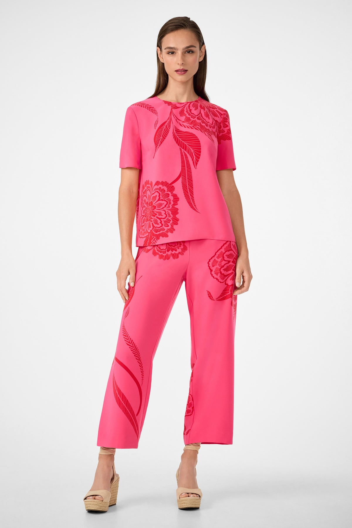 A woman stands against a plain white background wearing the Statue Pant—wide-leg, bright pink with bold red floral patterns—paired with a matching short-sleeve top and beige wedge sandals.
