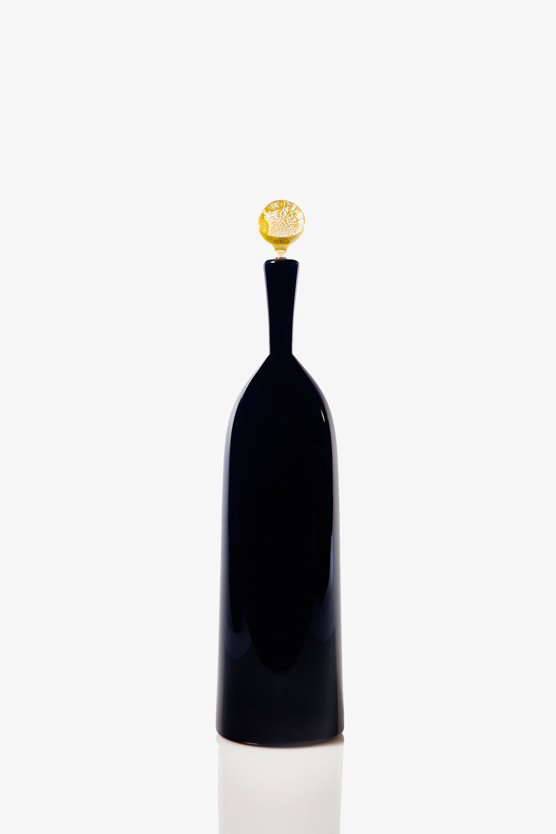 The Tall Bottle Carmella Barware features a sleek black glass body with a narrow neck and a round, textured golden stopper, displayed on a reflective white surface.