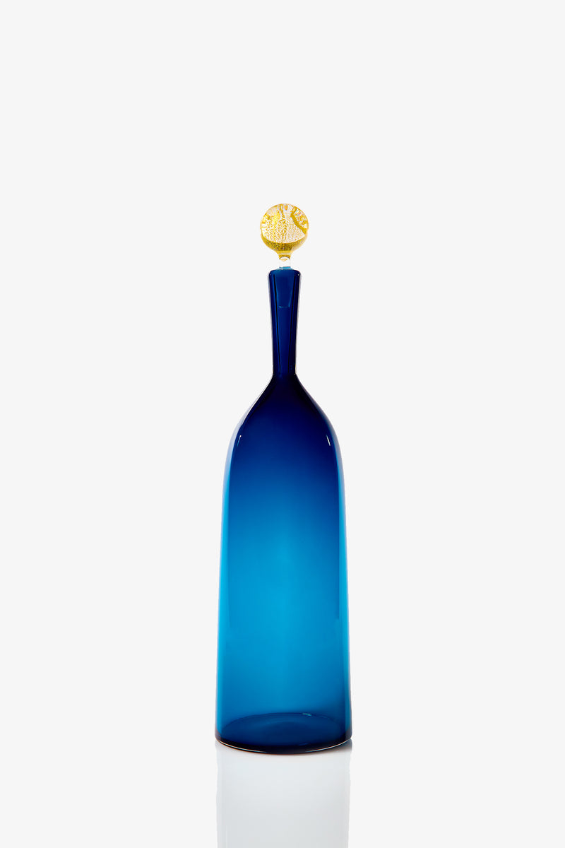 The Tall Bottle Carmella Barware features a slender blue glass design with a narrow neck and a round gold decorative stopper, displayed against a plain white background.