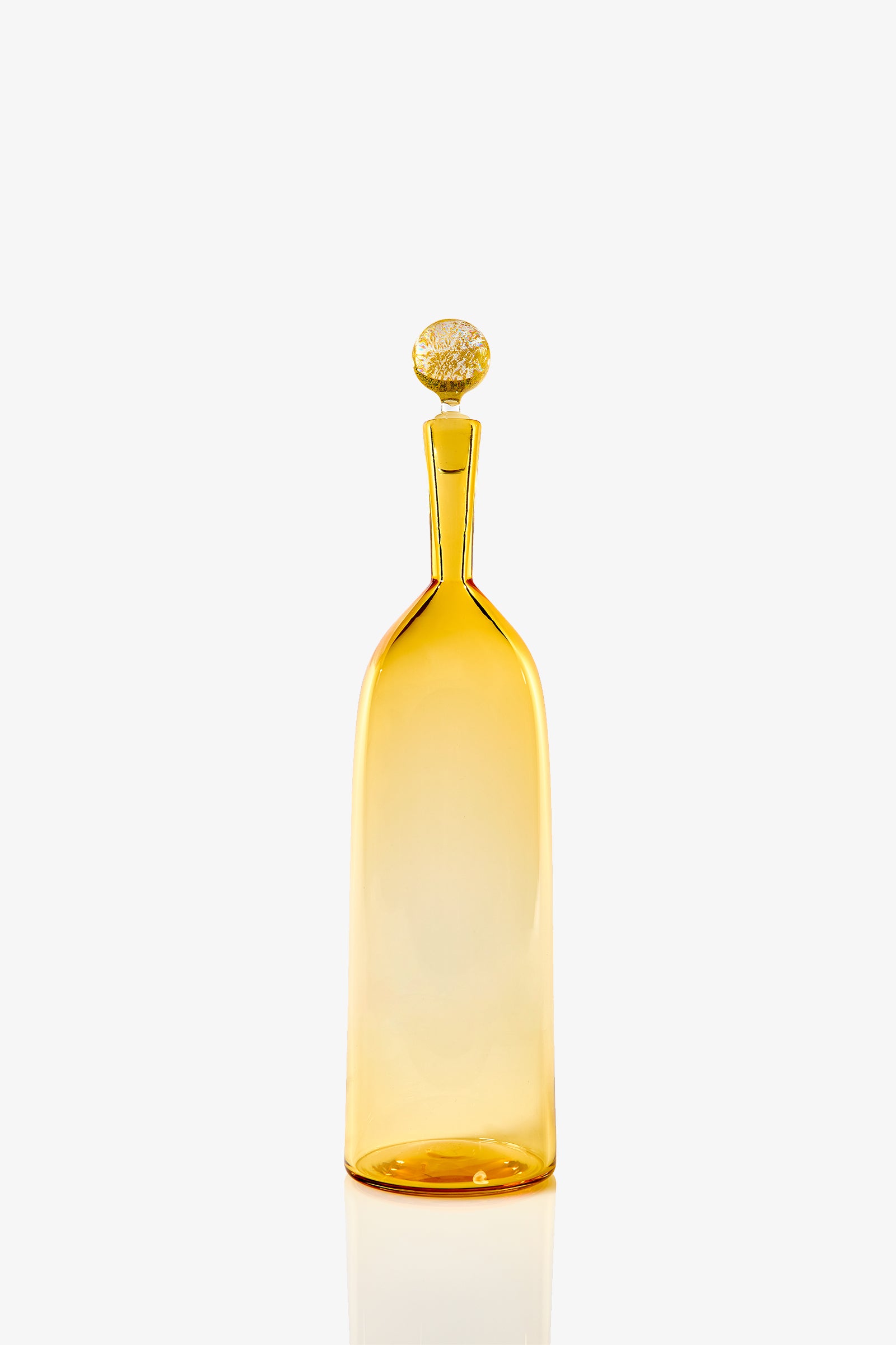 The Tall Bottle Carmella Barware is a slender amber glass bottle with a narrow neck and round, textured stopper, shown against a plain white background.