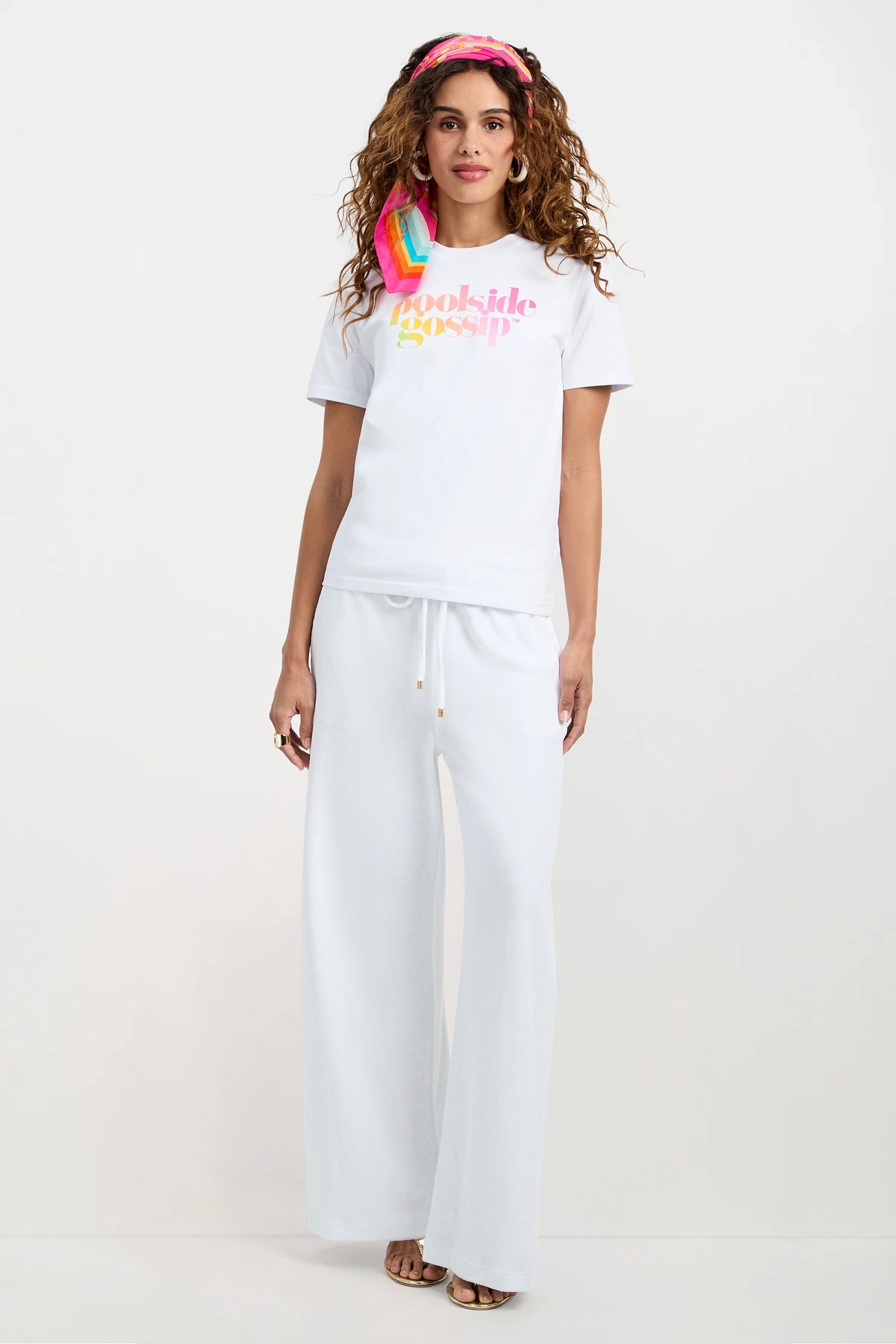 A woman with curly hair wears the Poolside Gossip Crewneck Souvenir Tee in white, paired with loose white pants and a pink headband. She stands barefoot against a plain white background, facing forward and smiling slightly.