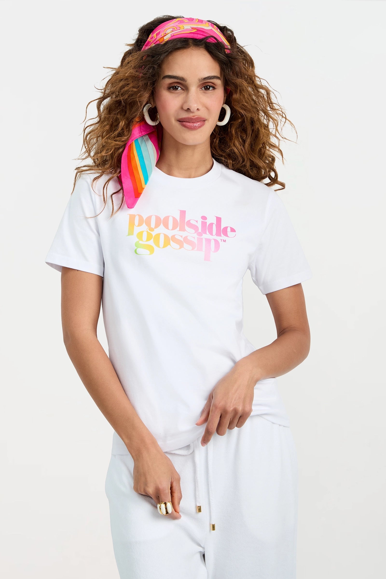 A woman with curly hair wearing a pink and yellow headscarf, white hoop earrings, and the Poolside Gossip Crewneck Souvenir Tee poses with a relaxed expression against a white background.