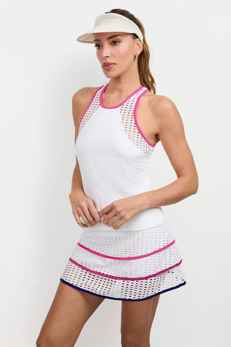 A woman in the Pointelle Trim Tank, featuring white fabric with pink details and mesh accents, paired with a striped skirt and beige visor, stands against a plain white background.