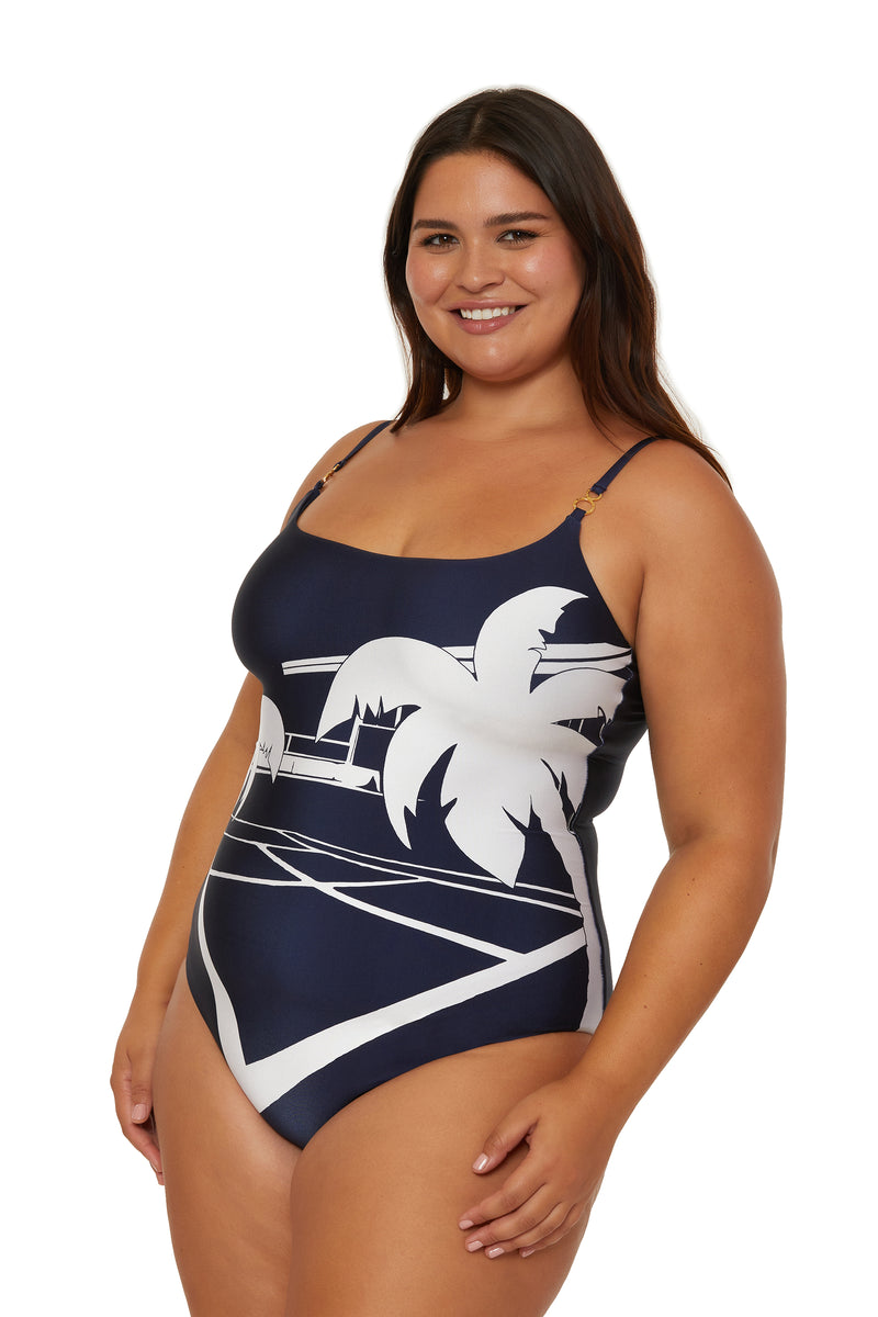 A smiling woman wears the Scoop Neck One-Piece Swimsuit - Trina Tennis, featuring a navy blue base with white palm tree and geometric designs, standing against a white background.