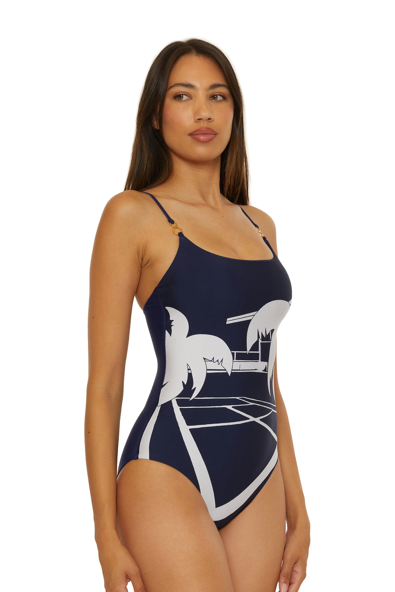 A woman models the Scoop Neck One-Piece Swimsuit - Trina Tennis, featuring a navy blue color with white abstract palm tree and beach graphics, thin straps, and a fitted design. She faces forward, looking slightly to her left.