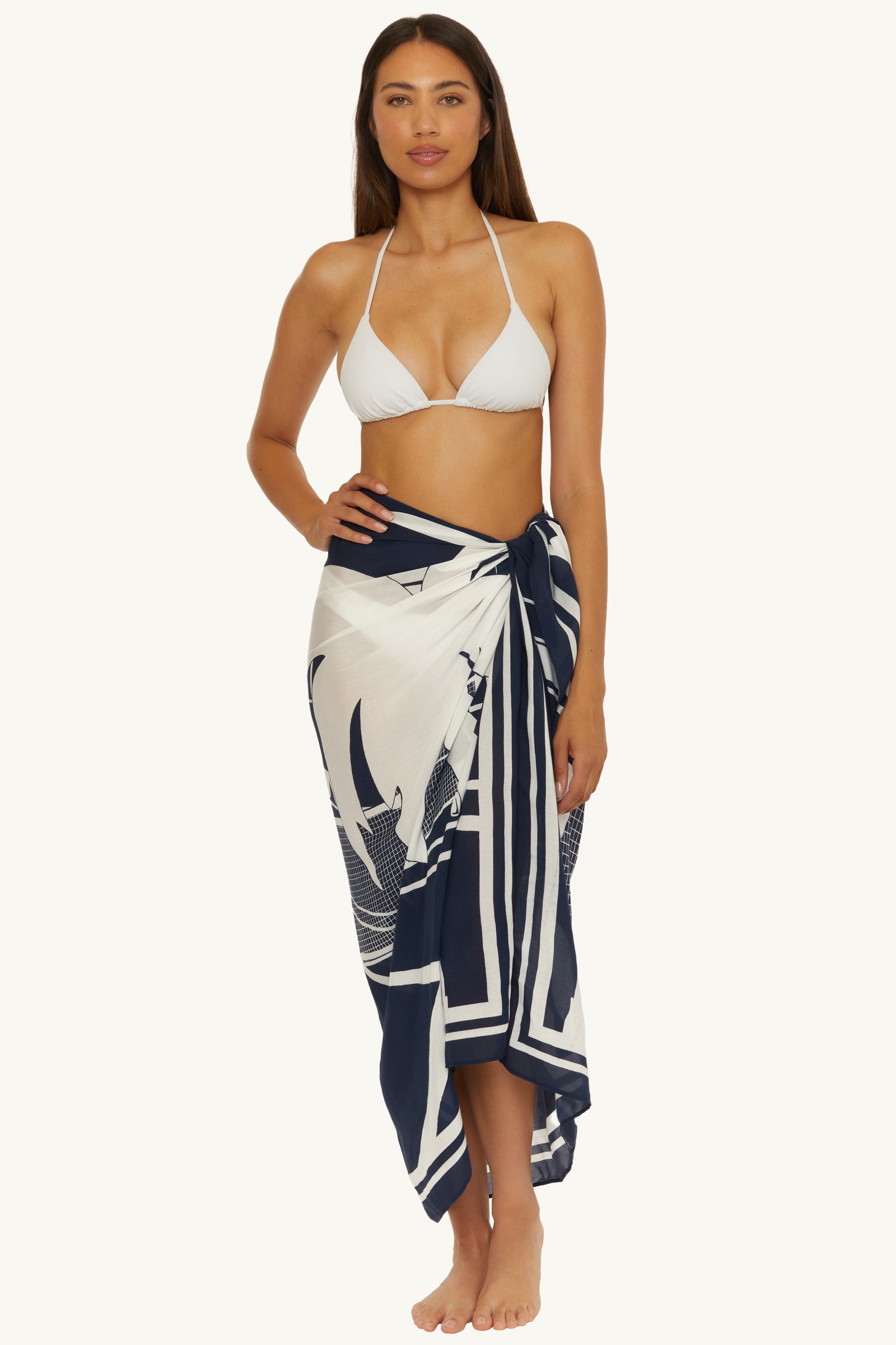 A woman stands barefoot in a white bikini top and the Pareo Swim Cover-Up - Trina Tennis in Ink, tied at the waist, against a neutral background.