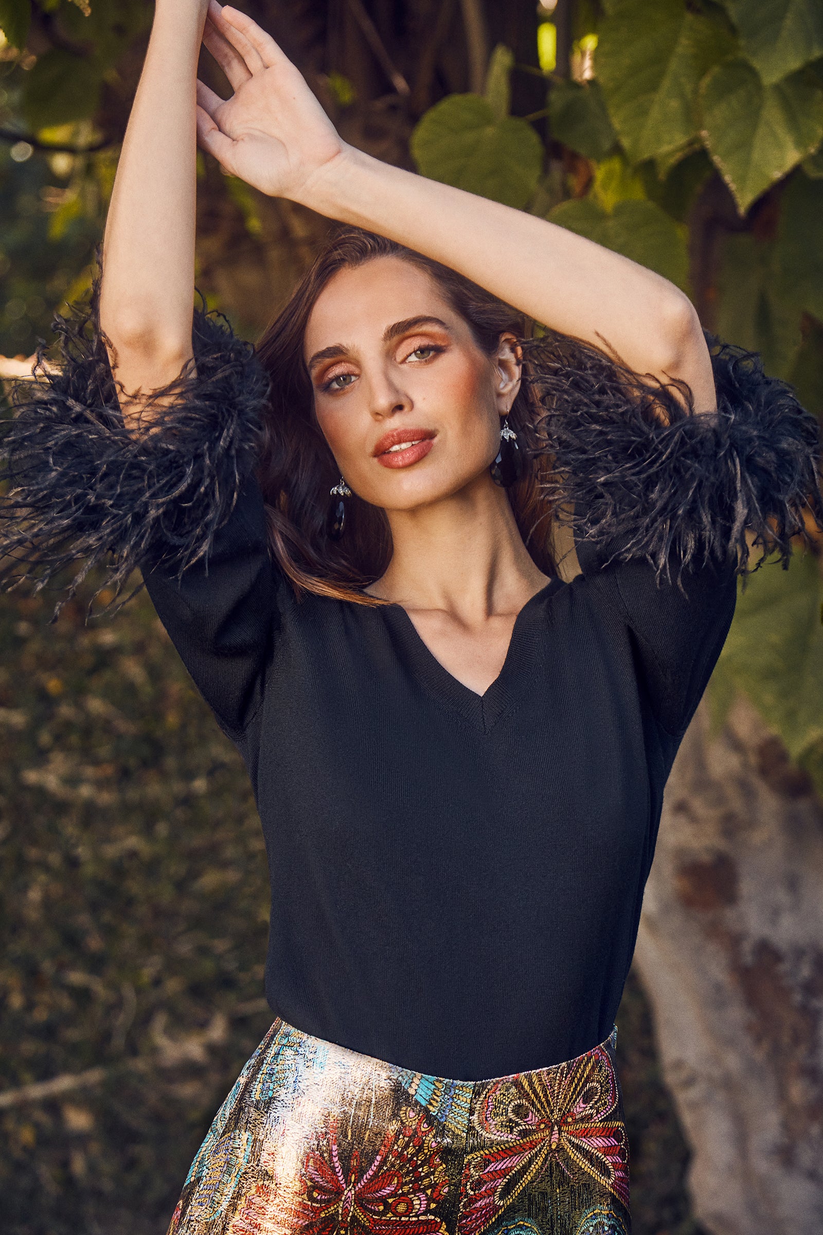 A woman poses outdoors with her arms raised, wearing the Sassy Merino Wool Feather Trimmed Sweater. Green leaves and natural sunlight frame the background.