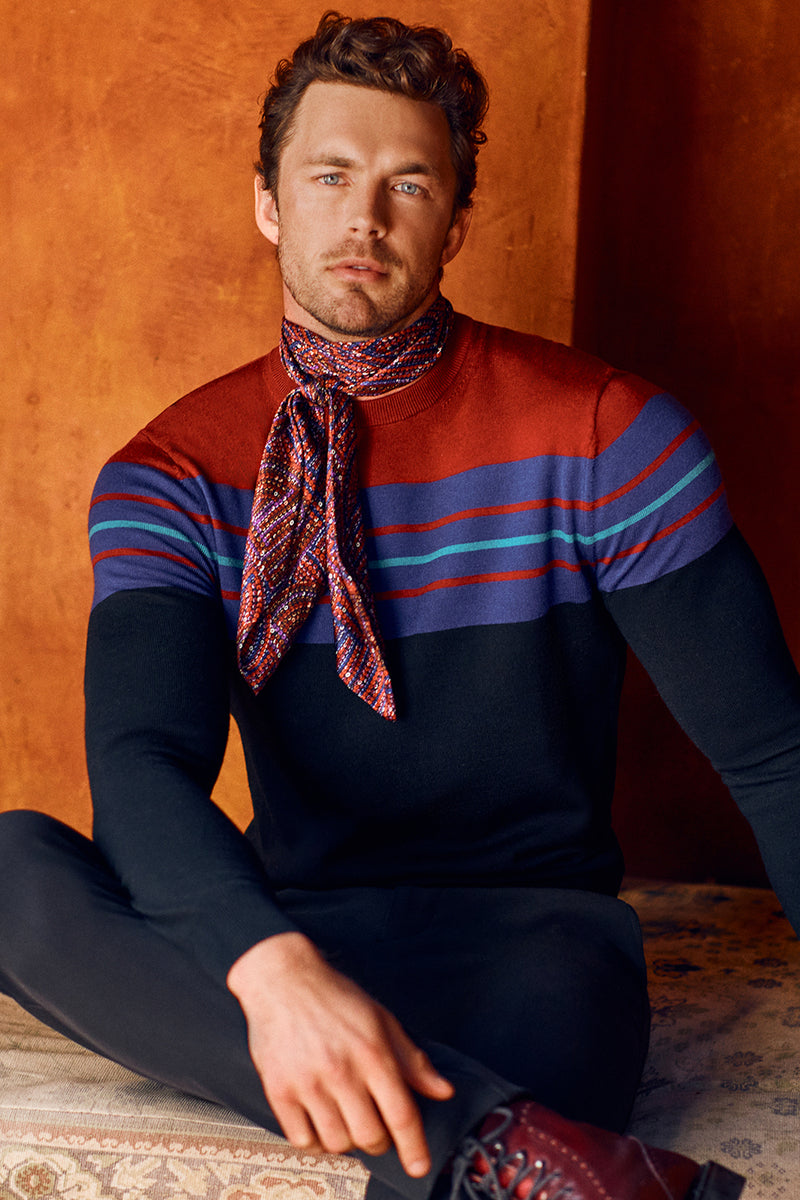 A man with short curly hair wears the Romero Crewneck Sweater, black pants, maroon boots, and a patterned scarf tied around his neck, sitting against an orange-brown textured background.