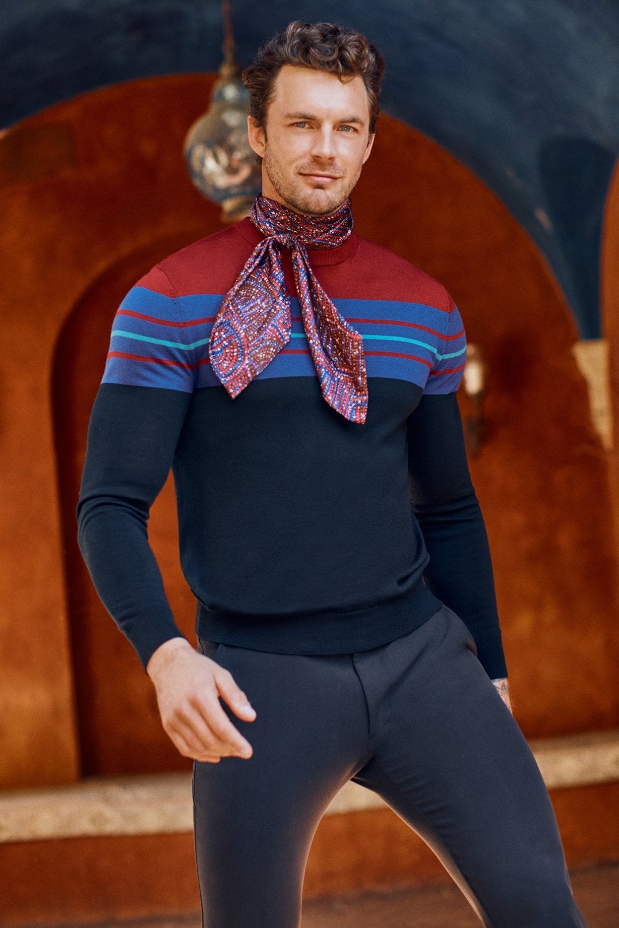 A man poses confidently against a warm terracotta backdrop, wearing the Romero Crewneck Sweater— a fitted, long-sleeve color-block design with horizontal stripes—paired with a patterned red scarf.