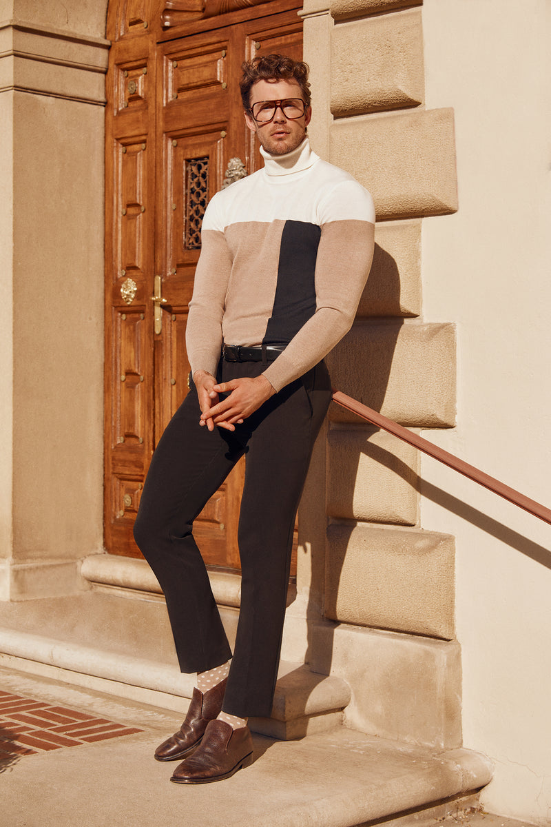 A man wearing glasses, black pants, brown shoes, and the Callisto Turtleneck Sweater - Merino Wool stands on stairs by a wooden door, posing with one hand on the railing.