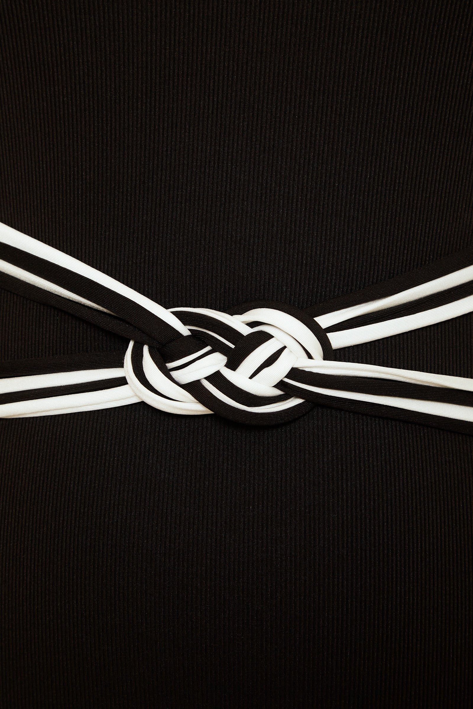 A close-up of the Triumph Braided One-Piece Swimsuit showcases two intertwined black and white striped fabric belts knotted at the center, forming a decorative pattern against its ribbed background.