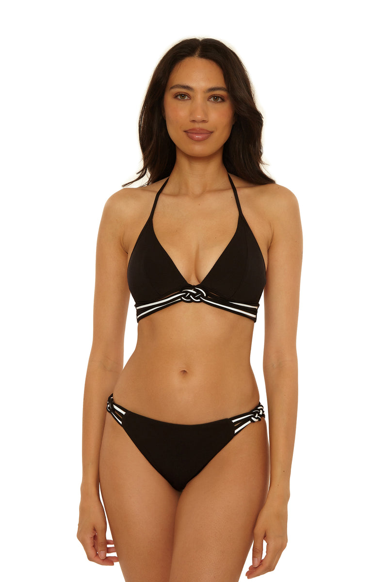 A woman with long dark hair and a neutral expression wears the Triumph Braided Hipster Swim Bottom, a black bikini featuring white striped trim and knot details on the top and sides of the bottoms.