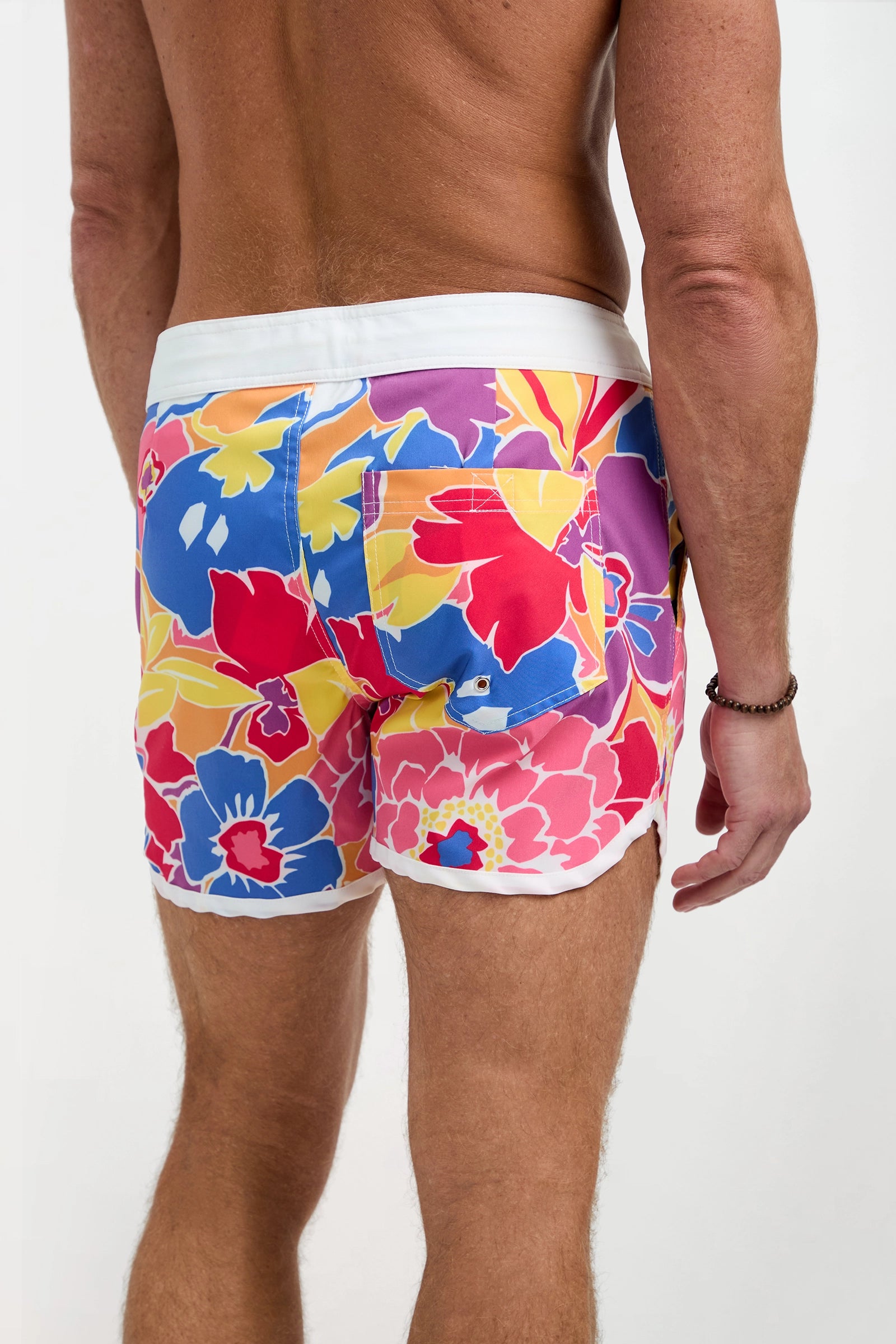 A man with a tan complexion stands with his back to the camera wearing the Surfside Board Short - Soie Paradis, featuring colorful floral patterns with red, yellow, blue, and pink flowers, a white waistband, and a black bracelet on his right wrist.