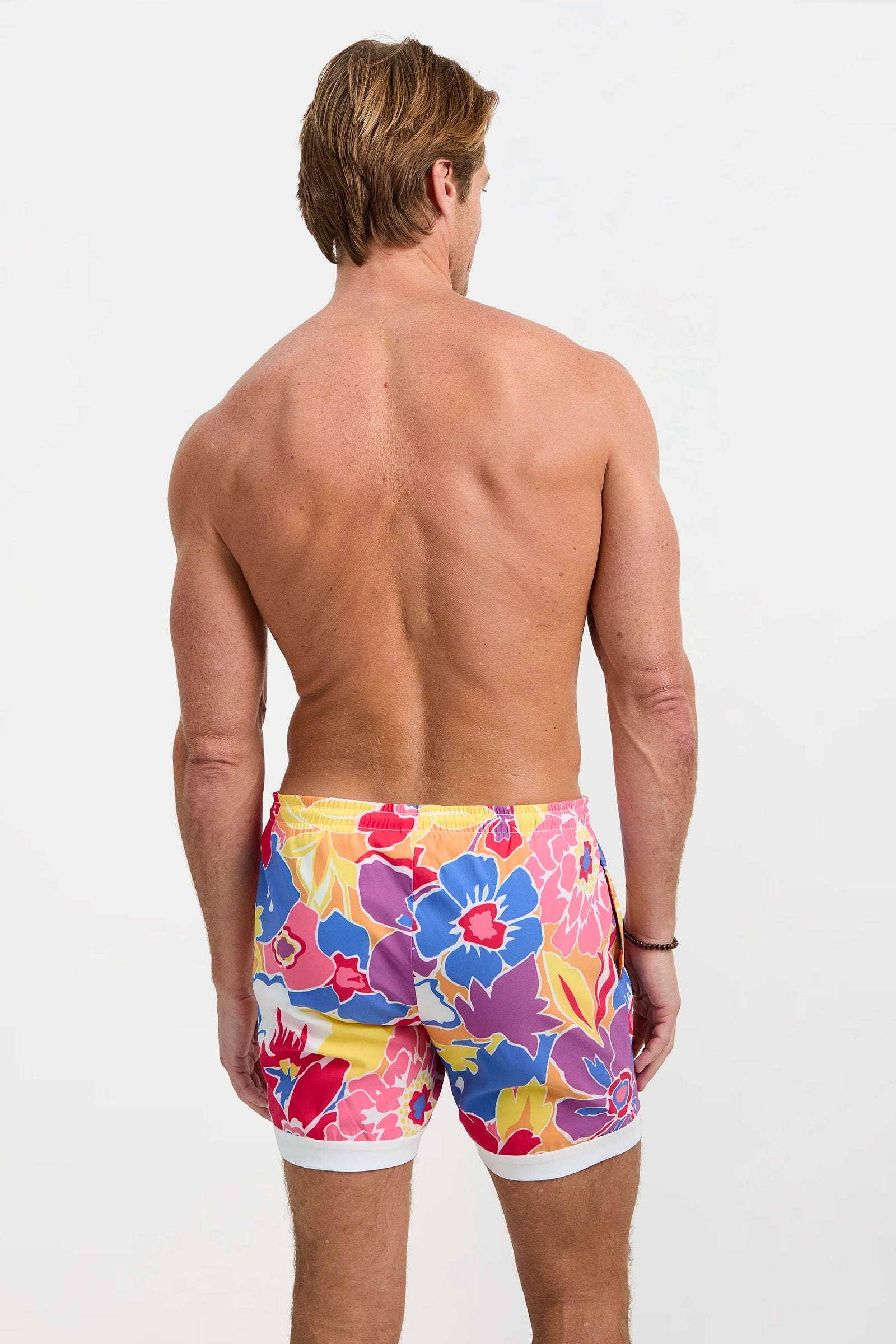 A shirtless man faces away wearing the Hermosa Beach Board Short - Soie Paradis, featuring a vibrant floral pattern in red, blue, yellow, and pink on a white background.
