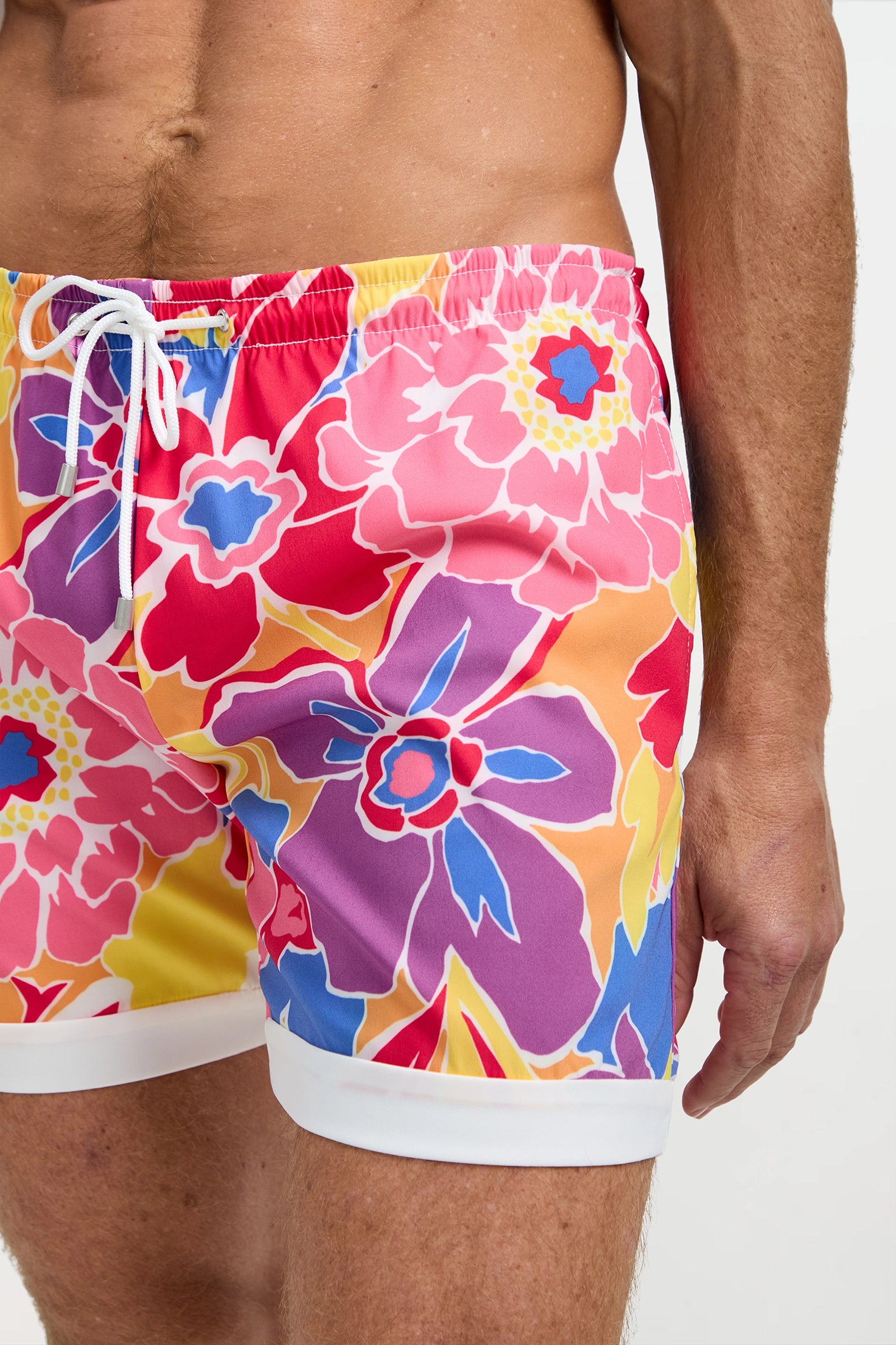 A close-up of a person wearing the Hermosa Beach Board Short - Soie Paradis, featuring a vibrant floral pattern in pink, purple, yellow, blue, and white with a white drawstring and trim.