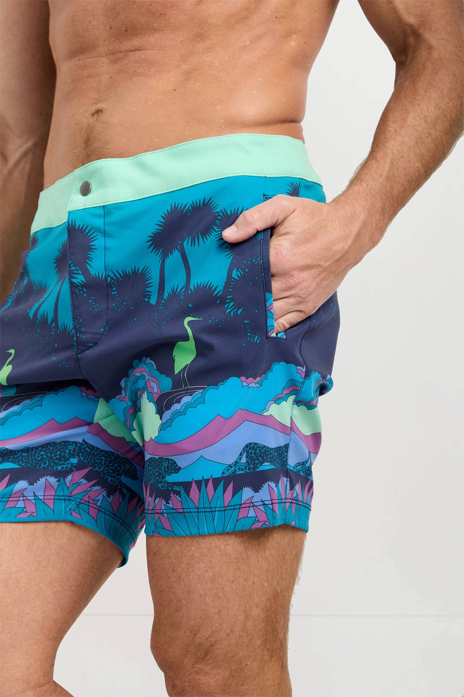 A man wears the Honolulu Board Short - Jungle, featuring a blue, purple, green, and teal jungle swim print, standing with one hand in his pocket against a plain background.