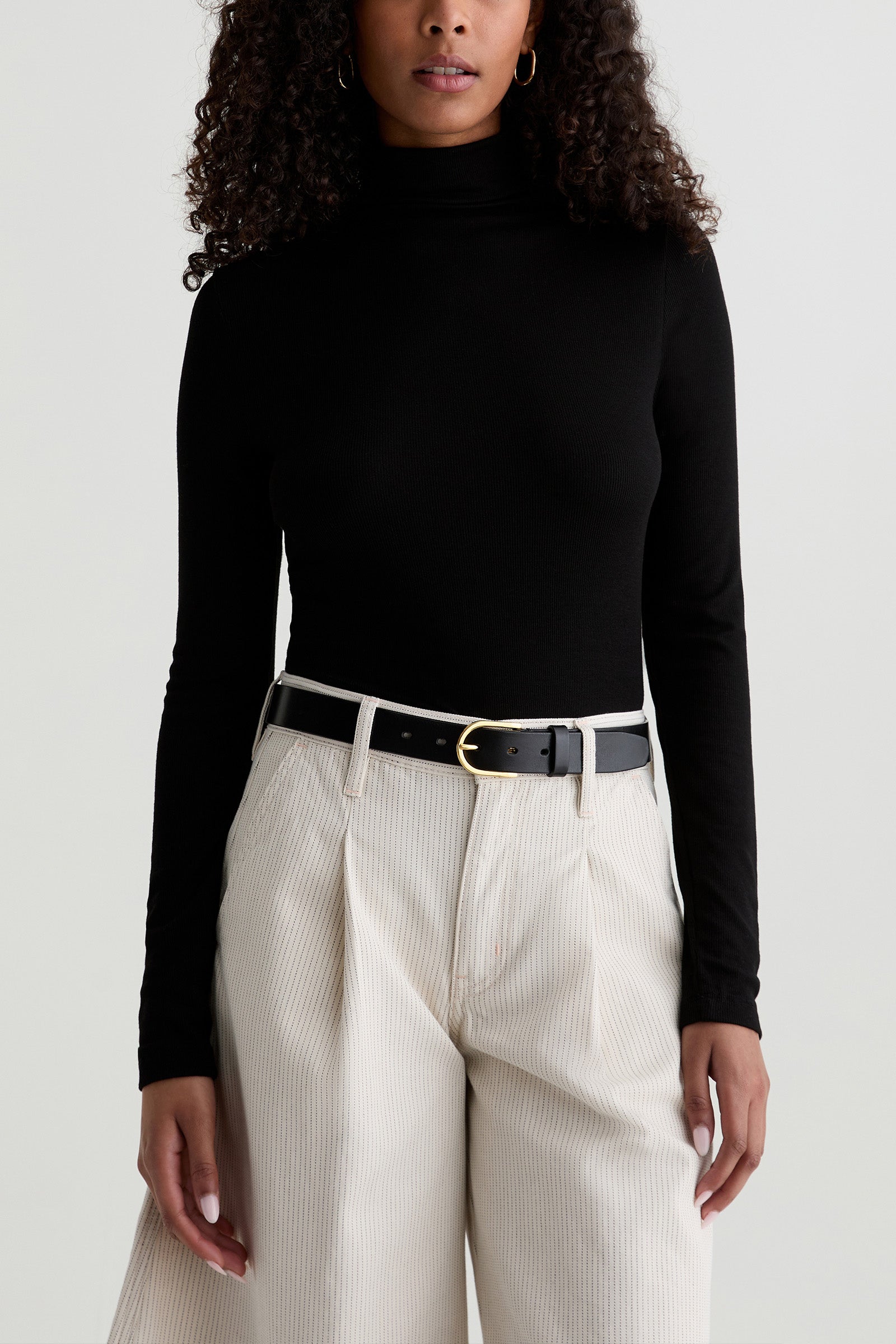A woman in the black AG Chelse Turtleneck, cream wide-leg pants, and a black belt with a gold buckle stands against a plain light background; her face is partially out of frame.