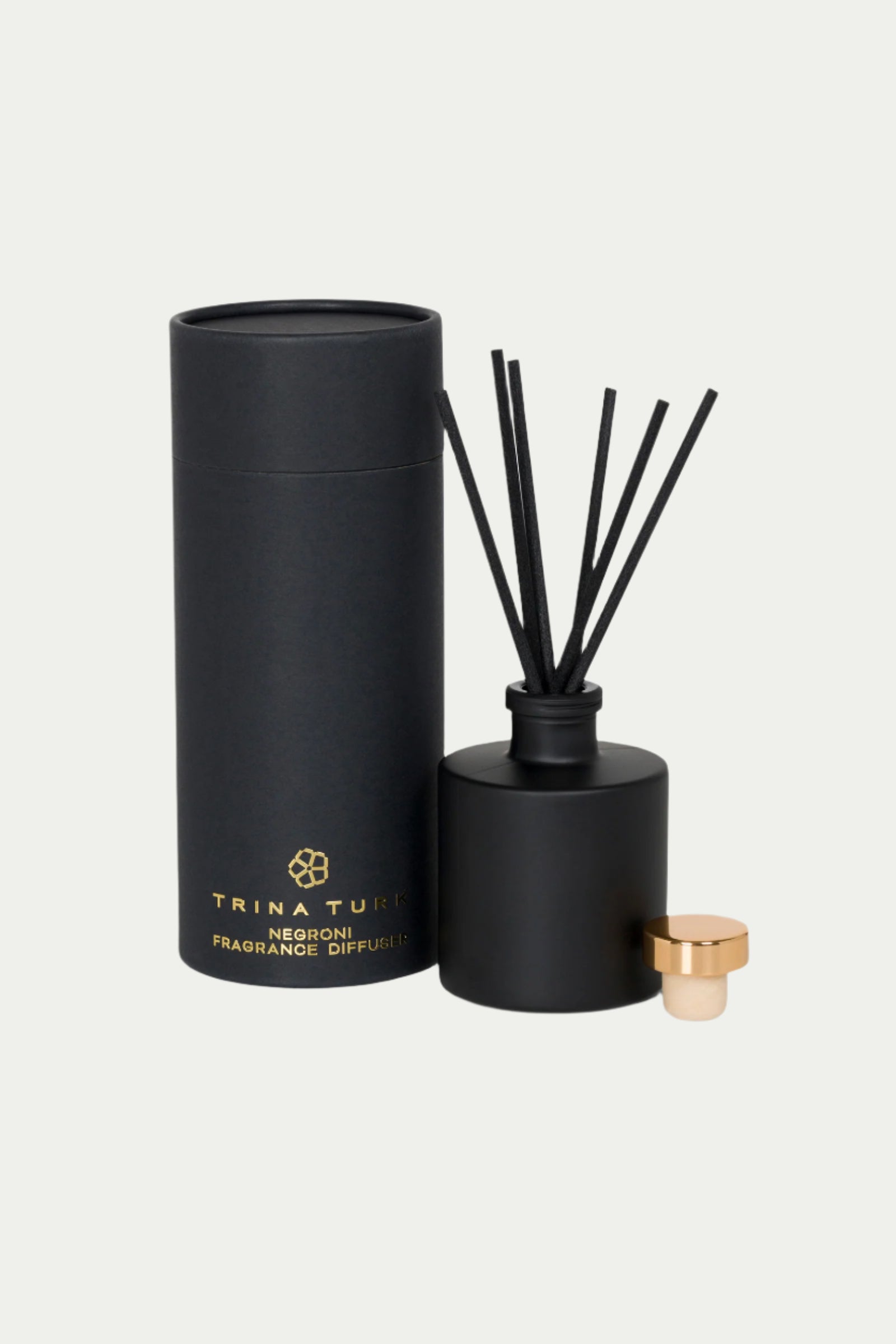 The TT x Perch Negroni Fragrance Diffuser features a sleek black cylindrical design with matching black reeds, alongside its coordinating black packaging adorned with gold lettering and logo. A small gold cap is placed near the diffuser.