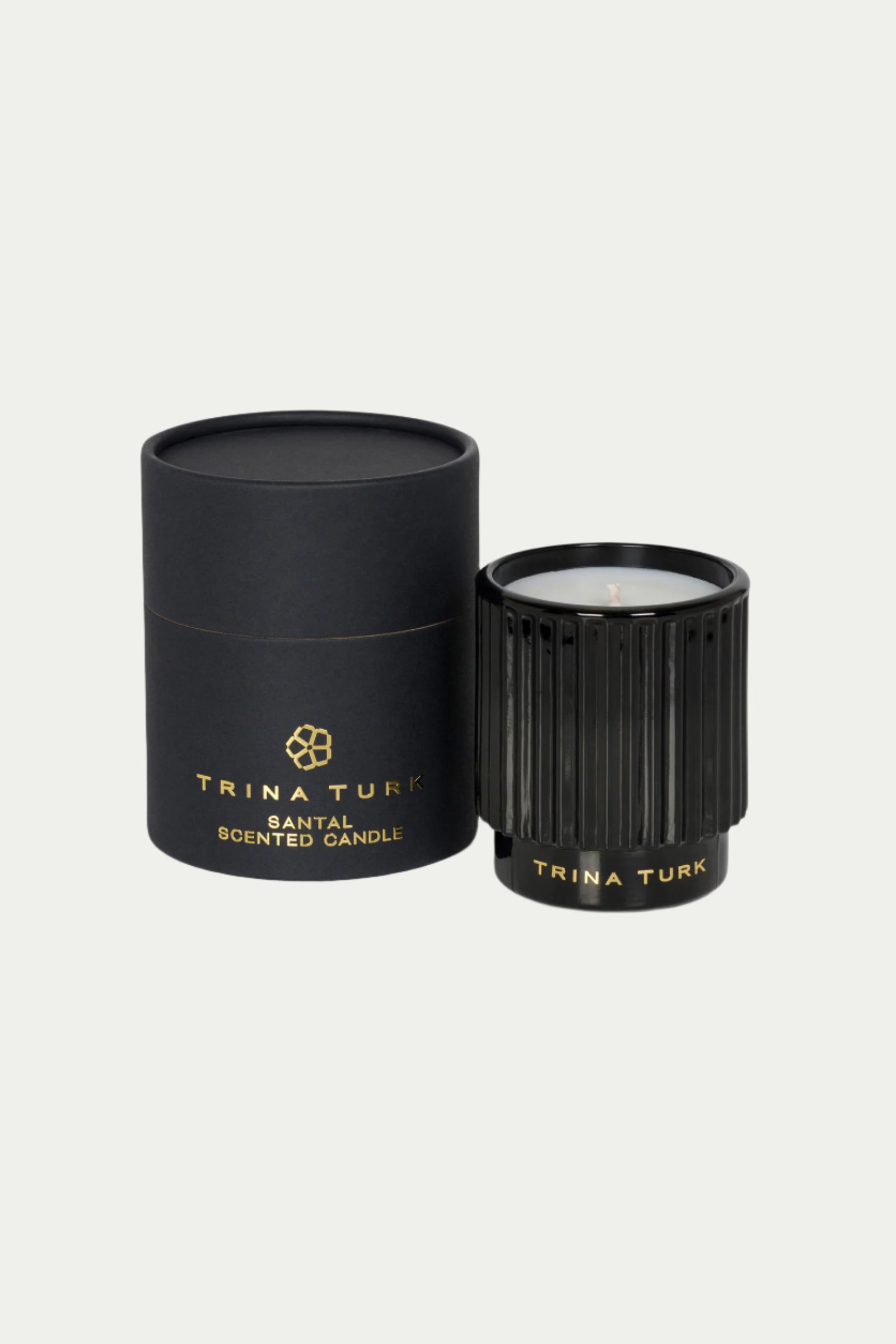 A black TT x Perch Santal Candle in a ribbed glass holder sits next to a matching black cylindrical box, both featuring gold lettering.